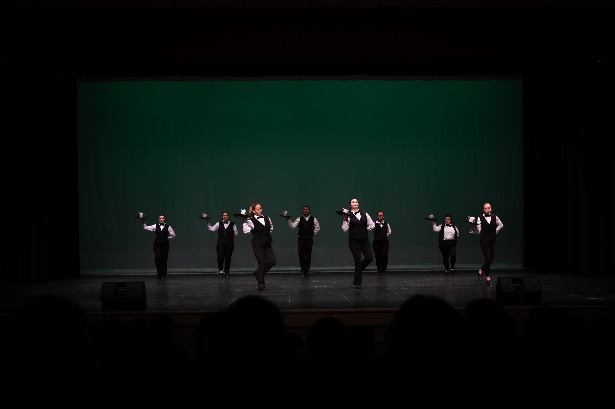 The image shows a group of performers on stage dressed in black vests and pants with white shirts, holding trays with teapots. They are performing a coordinated dance or routine against a green stage background with an audience in front.