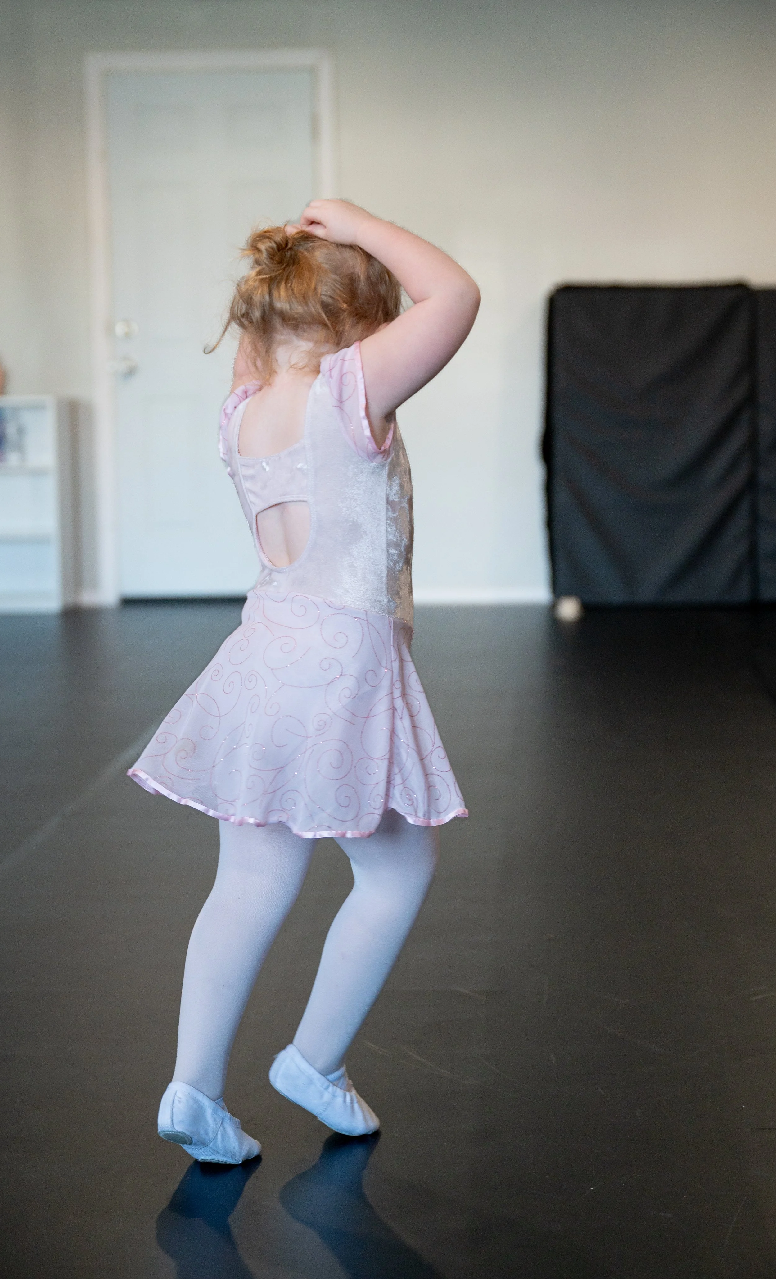 A young girl with red hair wearing a pink ballet dress, white tights, and ballet slippers, dancing on a black floor in a room with a white door and black furniture in the background.