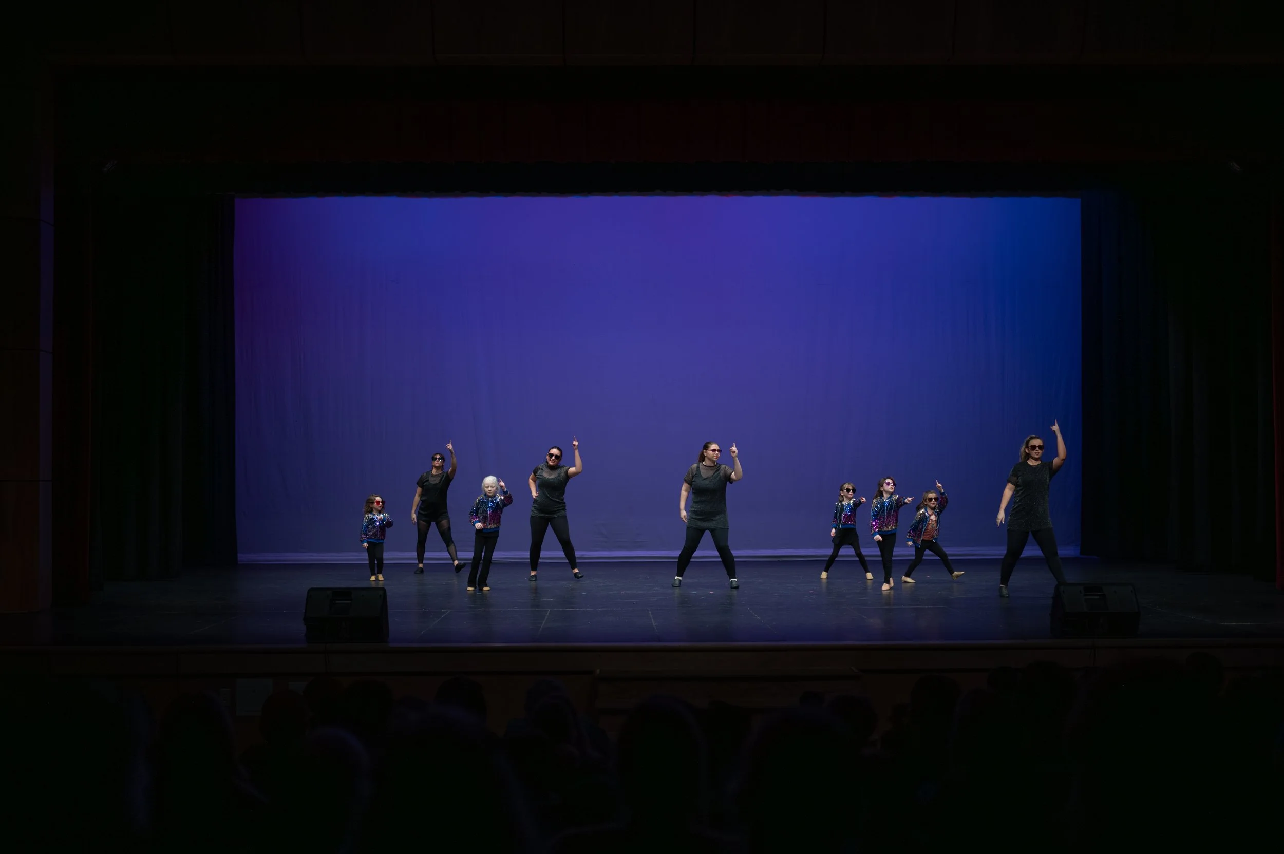 Group of children and adults performing a dance on stage with blue background.