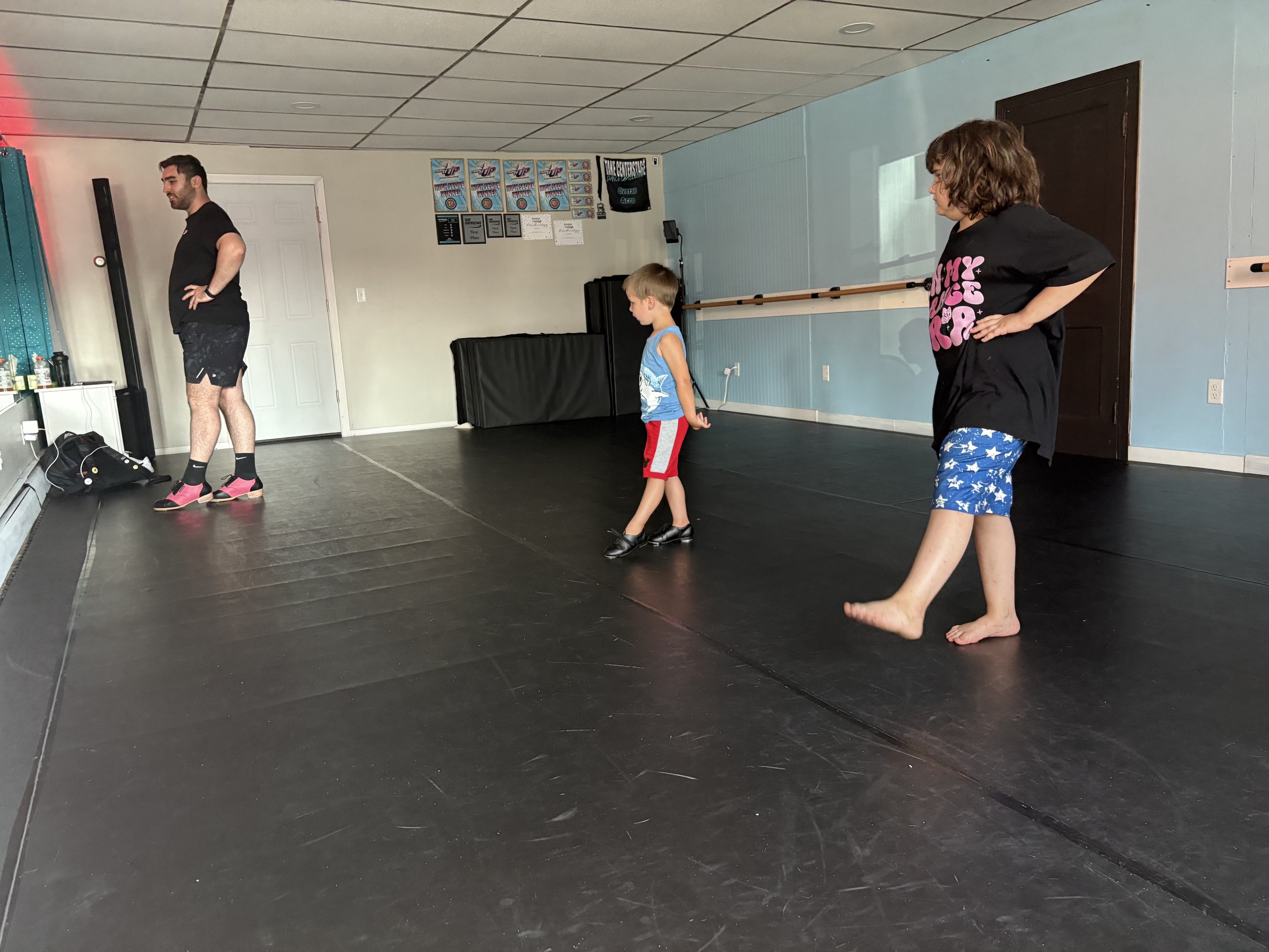 A ballet or dance studio with three people: an adult male on the left wearing black clothing and pink shoes, a young boy in the center wearing a blue tank top with white design and red shorts, and an adult woman on the right wearing a black t-shirt and blue star-patterned shorts. All are standing on a black dance floor.
