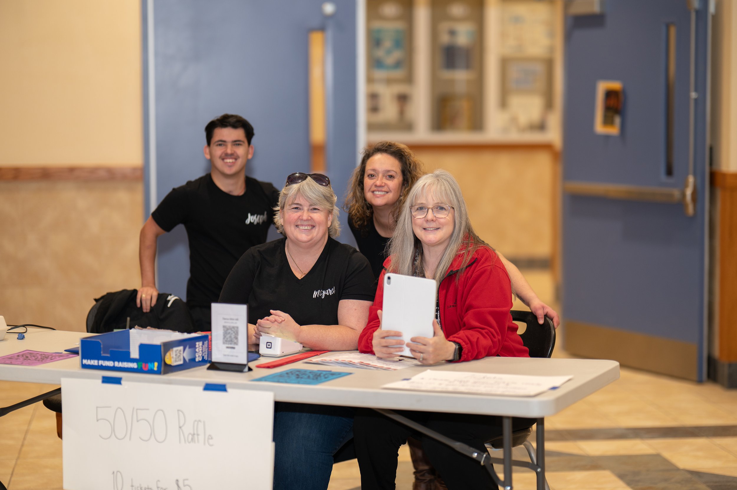 Four smiling women posing at a fundraising event with a raffle sign on the table. One woman is holding a tablet, and there are raffle tickets and a QR code on the table. The background shows a blue door and a display of pictures on the wall.