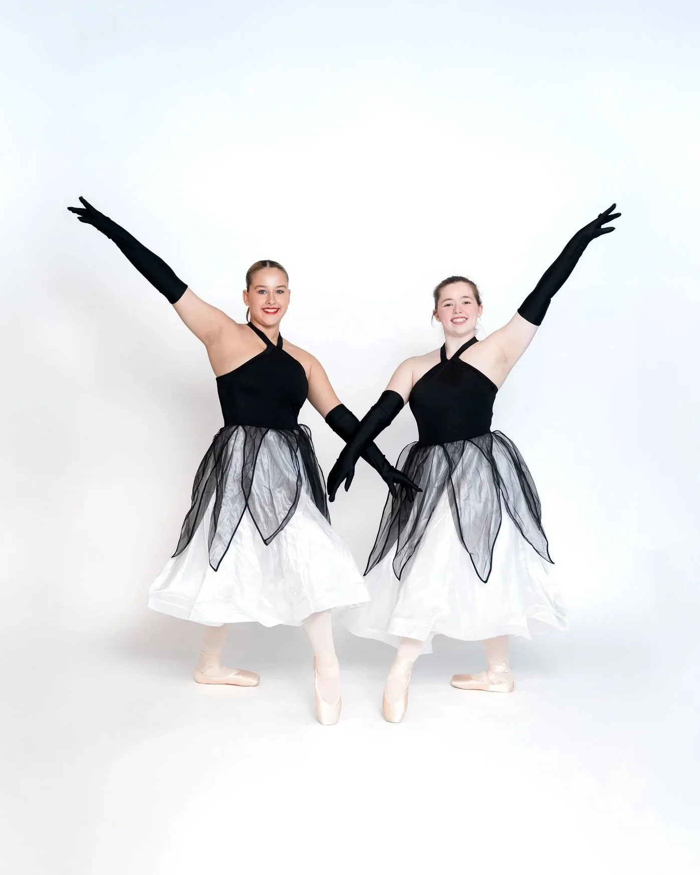 Two ballet dancers in black and white costumes with black gloves, posing against a white background, standing on pointe with arms raised.