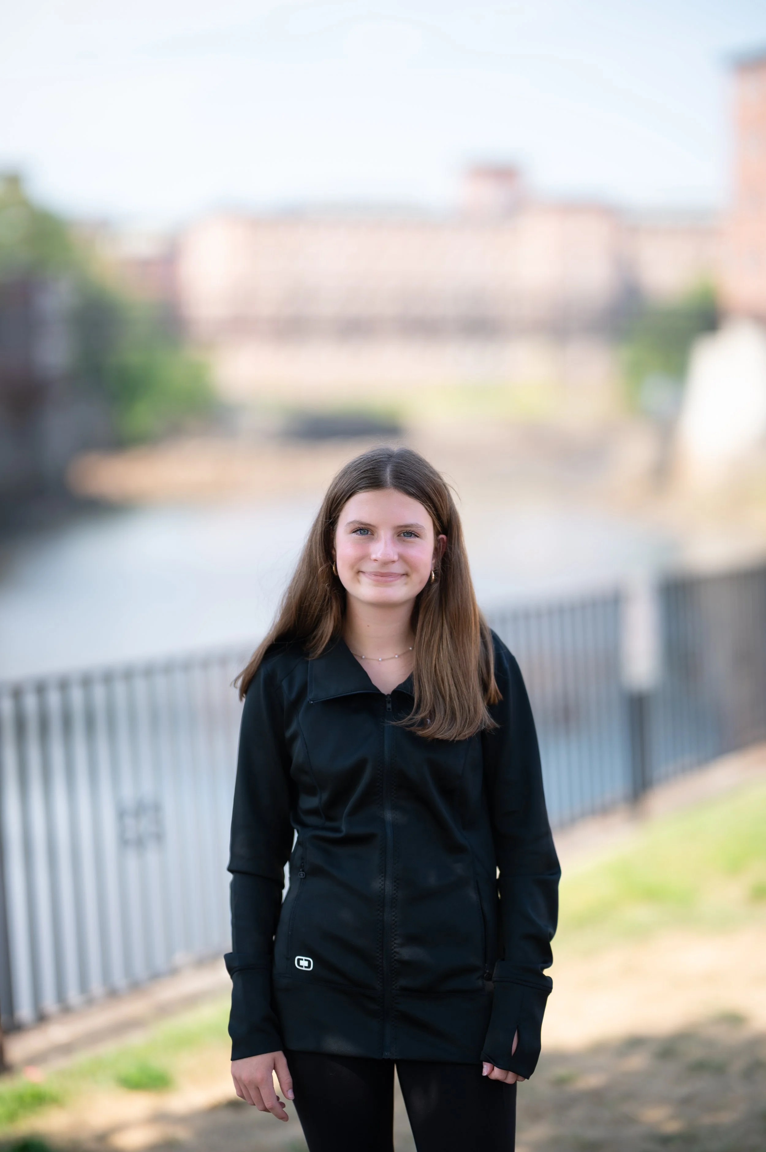 A teenage girl with long brown hair and a black zip-up jacket standing outdoors near a river, with a metal railing behind her and buildings in the background.