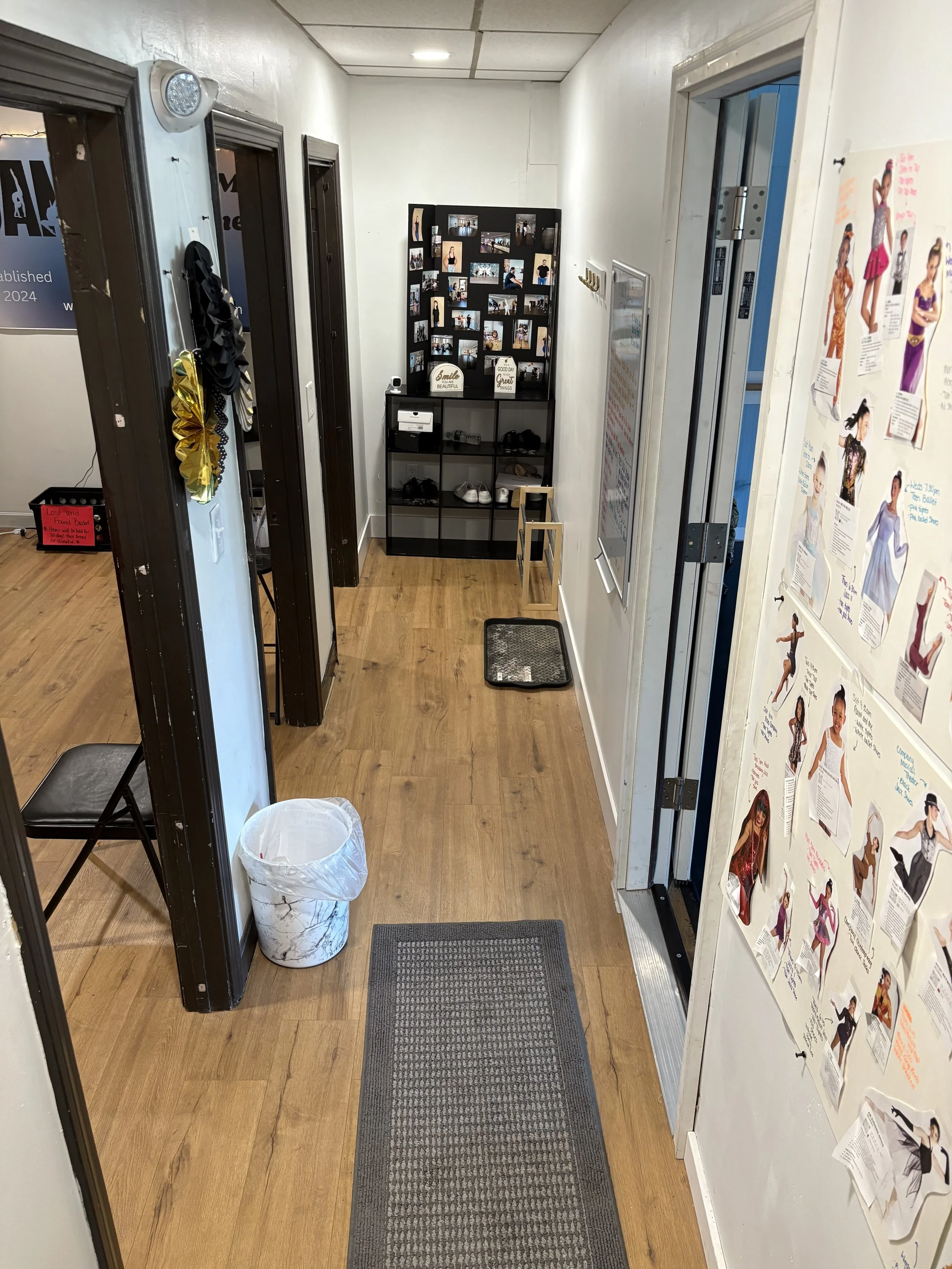 Hallway with wooden floor, black-framed doors, a white trash bin, a small black chair, and a shoe rack at the end. On the right wall, a whiteboard and a colorful poster featuring various women and dance poses.