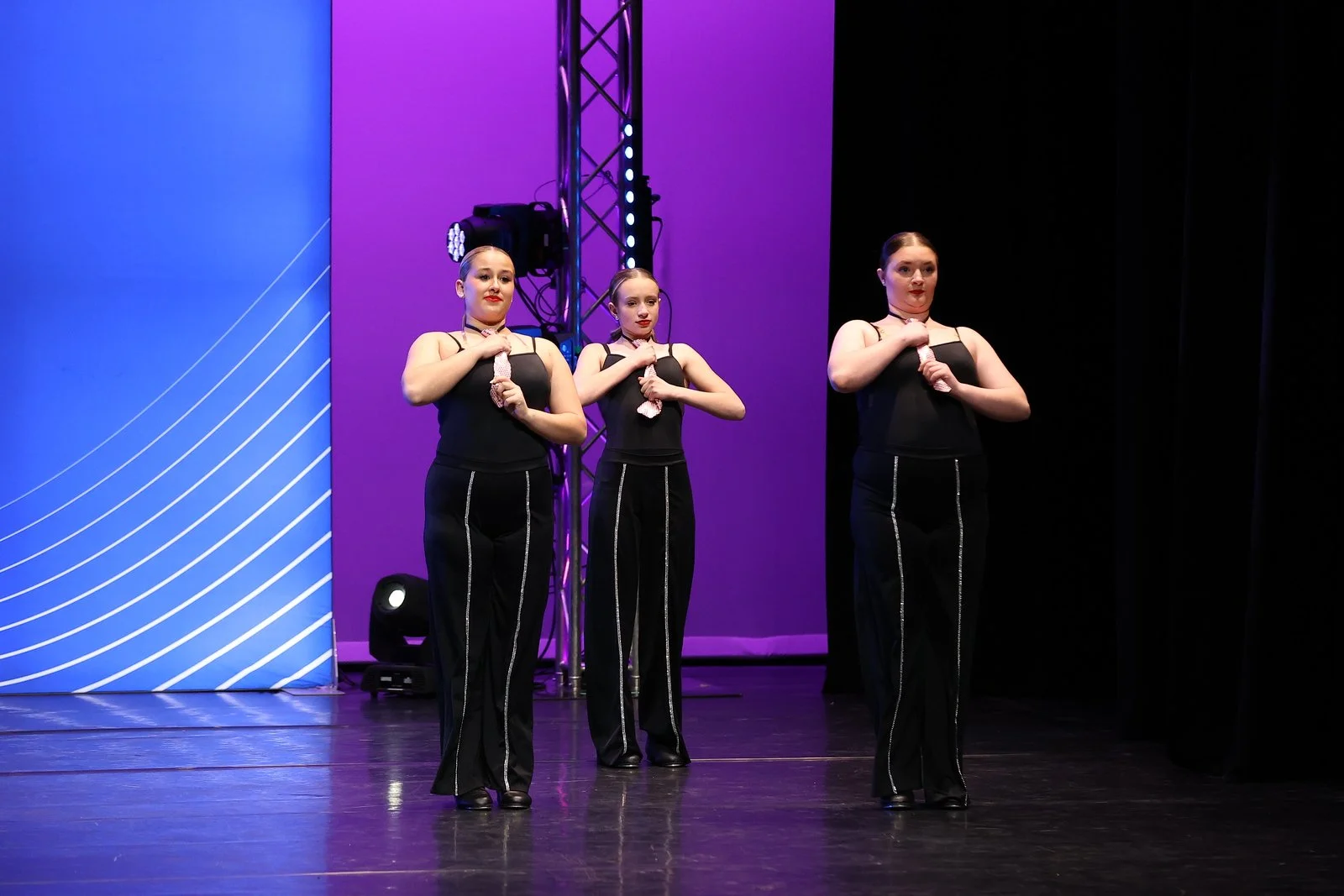 Three young women in black costumes performing a dance or routine on stage, standing with hands over their chests, in front of a colorful lit background.
