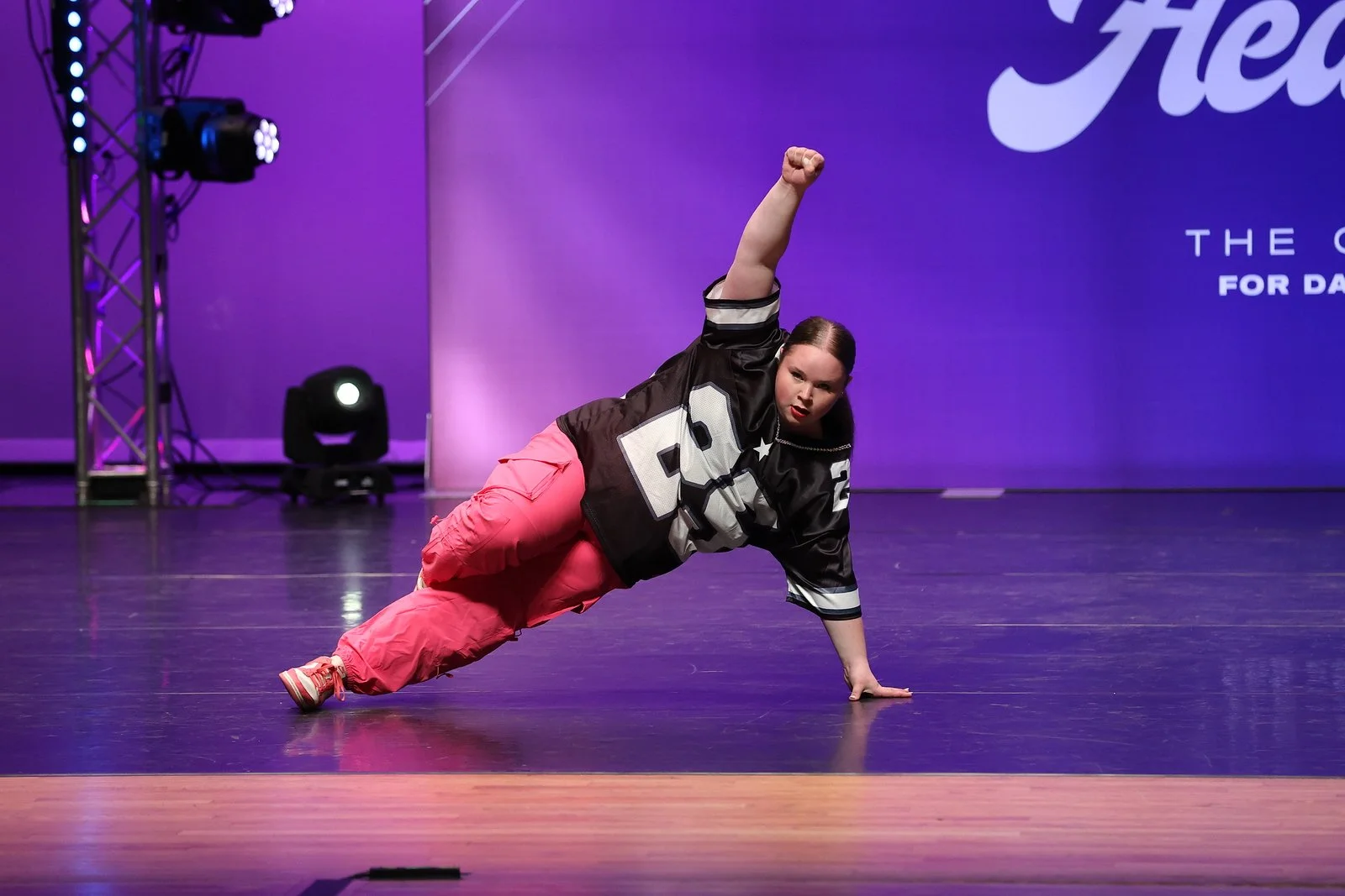 A female dancer performing a dance move on stage, wearing a black varsity jacket and pink pants, with one arm raised and the other hand on the floor.