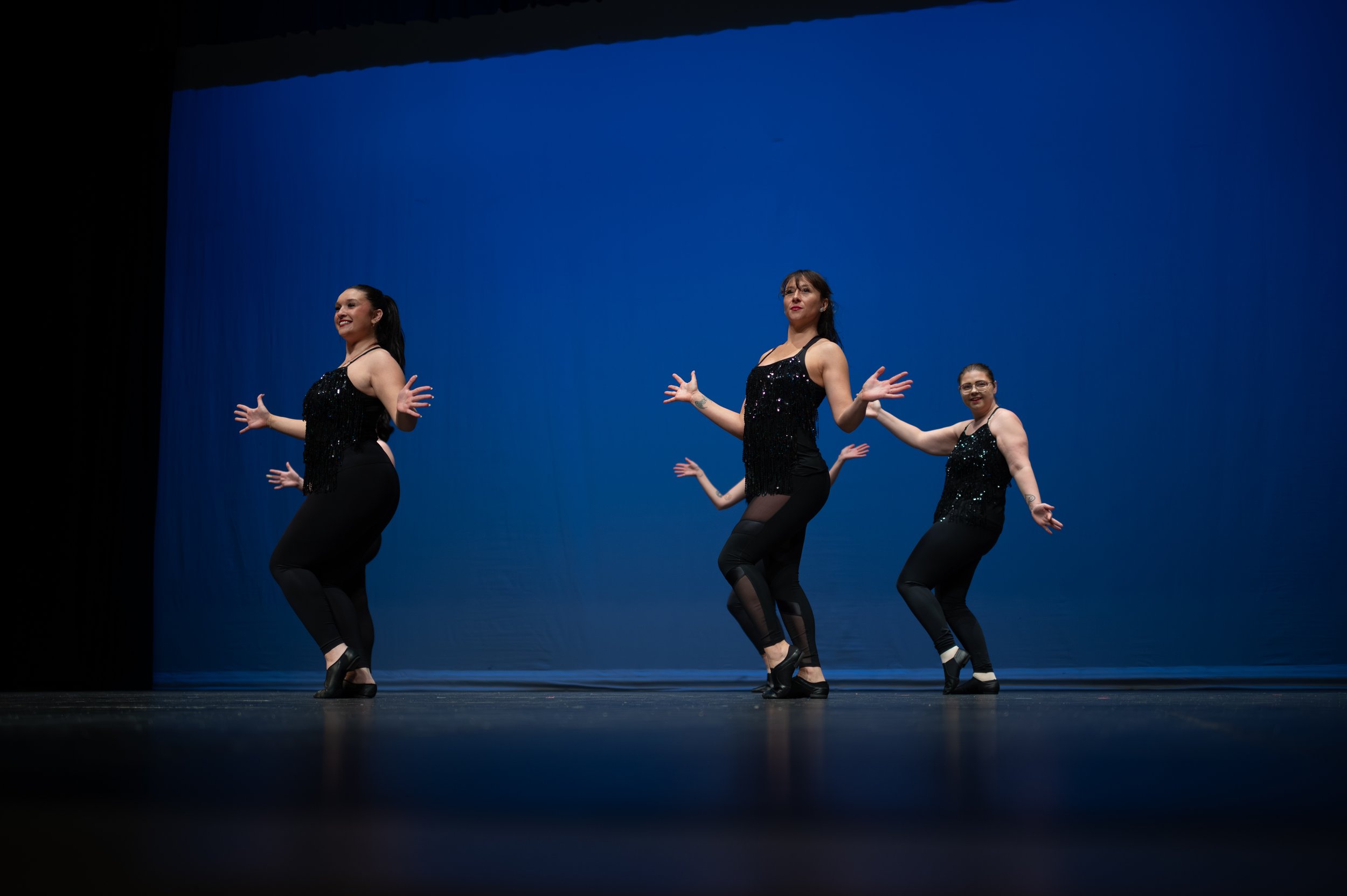 Four women in black sequin tops and black pants perform a dance on a stage with a blue backdrop.