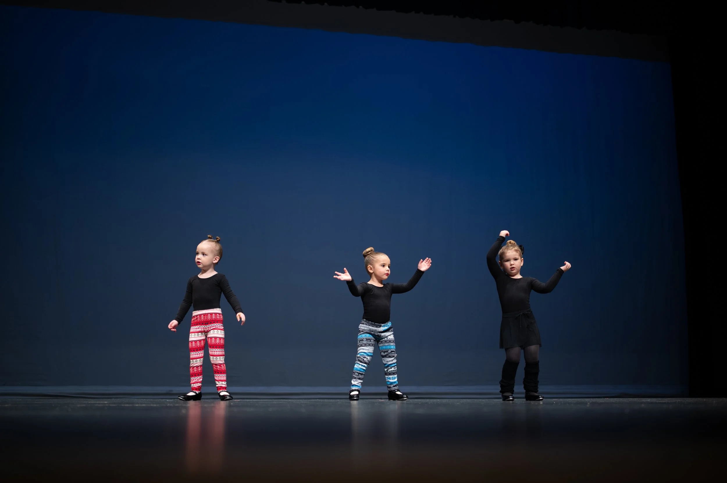 Three young girls perform a dance routine on stage, dressed in black tops and colorful patterned pants, with a dark blue backdrop.