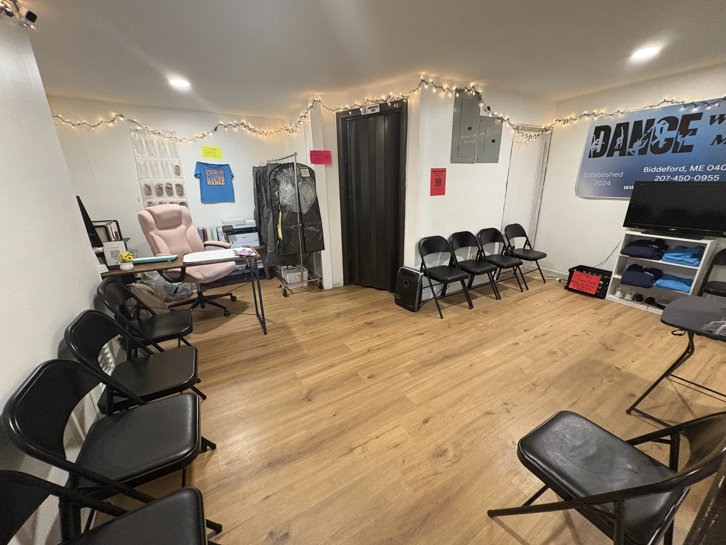 Dance With ME waiting room with chairs arranged along the walls, a desk with a pink chair, a banner with 'DANCE' on the wall, string lights, and a TV on a shelf with folded clothes.