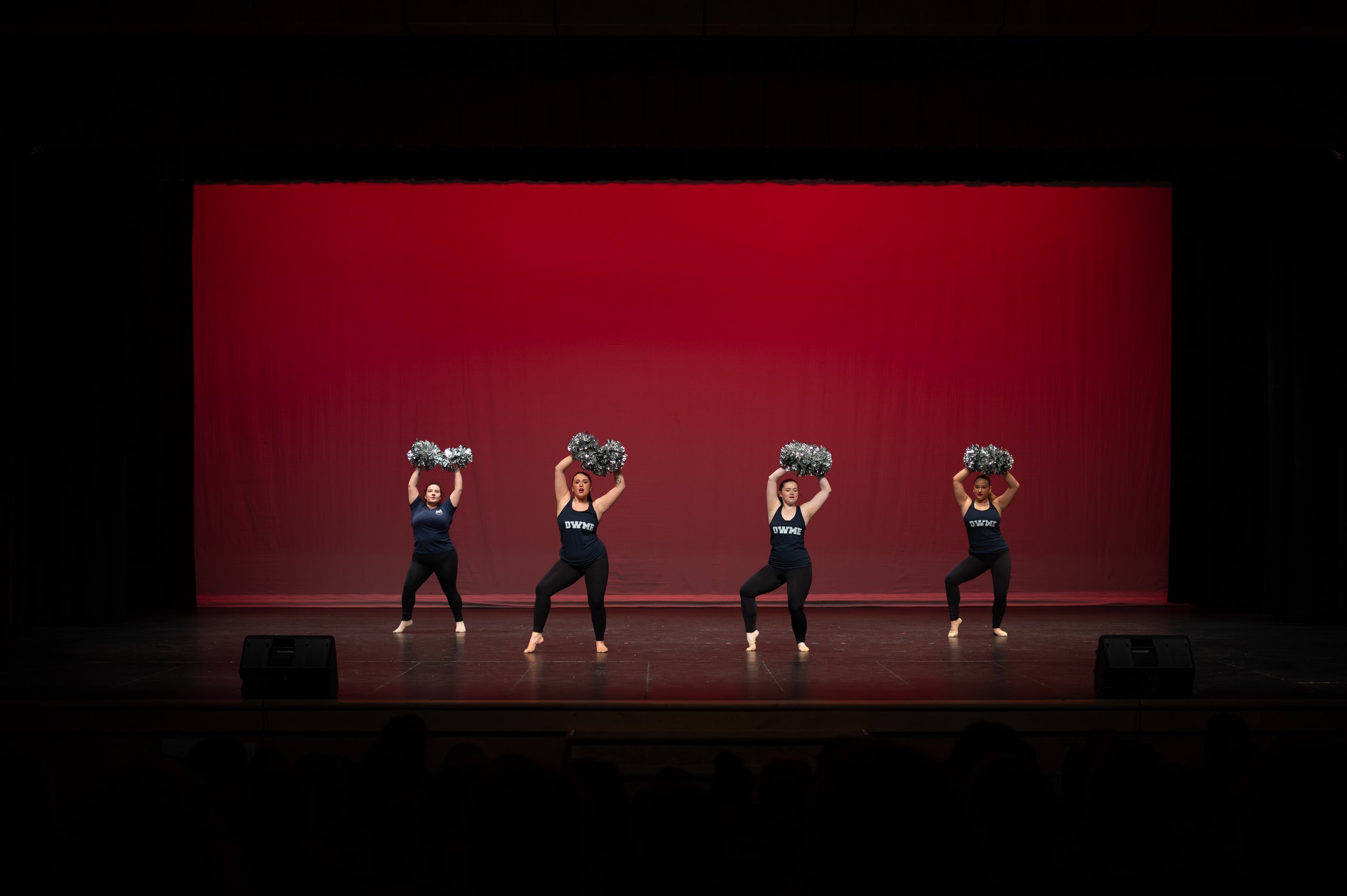 Four cheerleaders perform a routine on stage, holding silver pom-poms above their heads, with a red backdrop behind them.