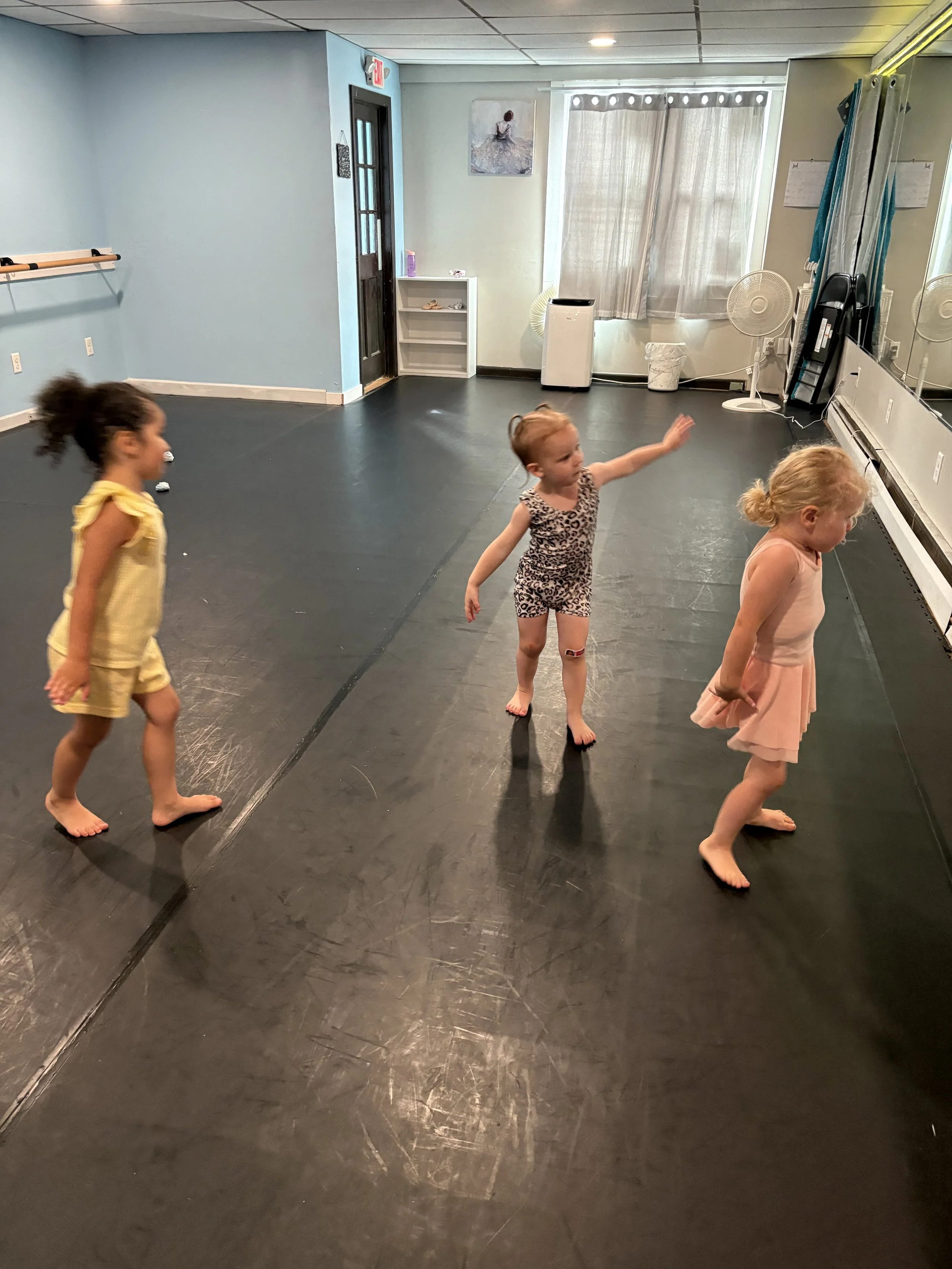 Three young girls dancing or playing in an indoor dance studio with a black floor, light blue walls, a music mirror wall, and a small artwork on the wall. All three are barefoot.