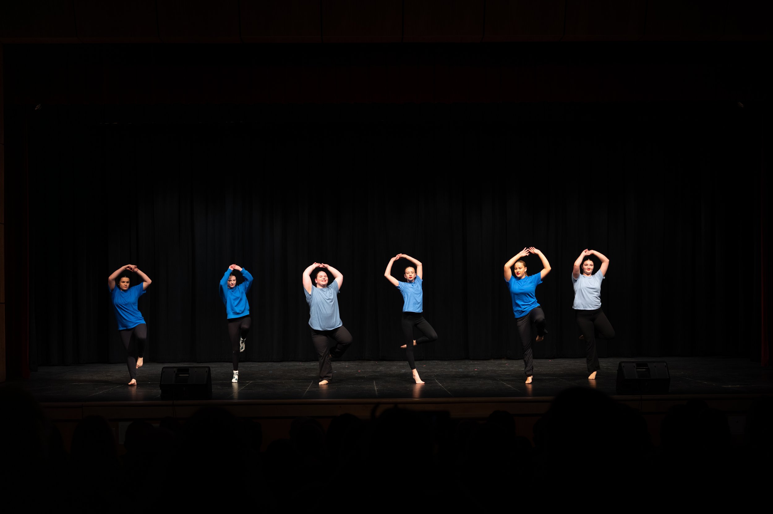 Six dancers on stage performing a routine, wearing blue tops and black pants, with a black curtain backdrop.
