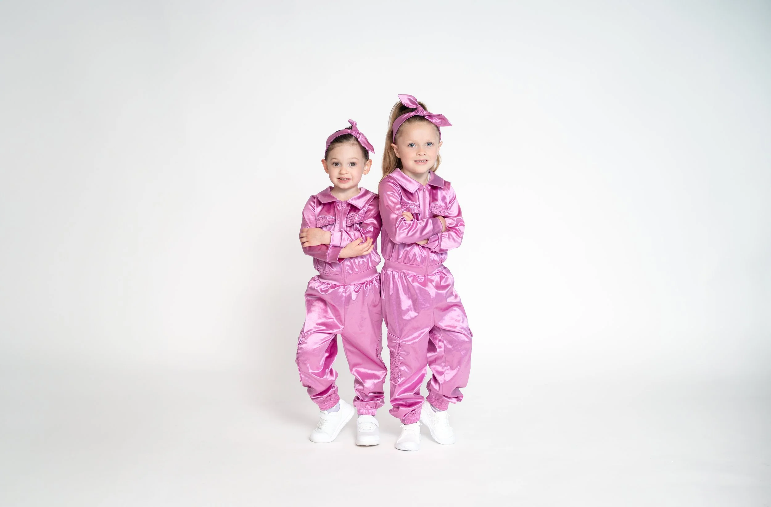 Two young girls in matching pink satin jumpsuits and headbands standing with arms crossed against a plain white background.