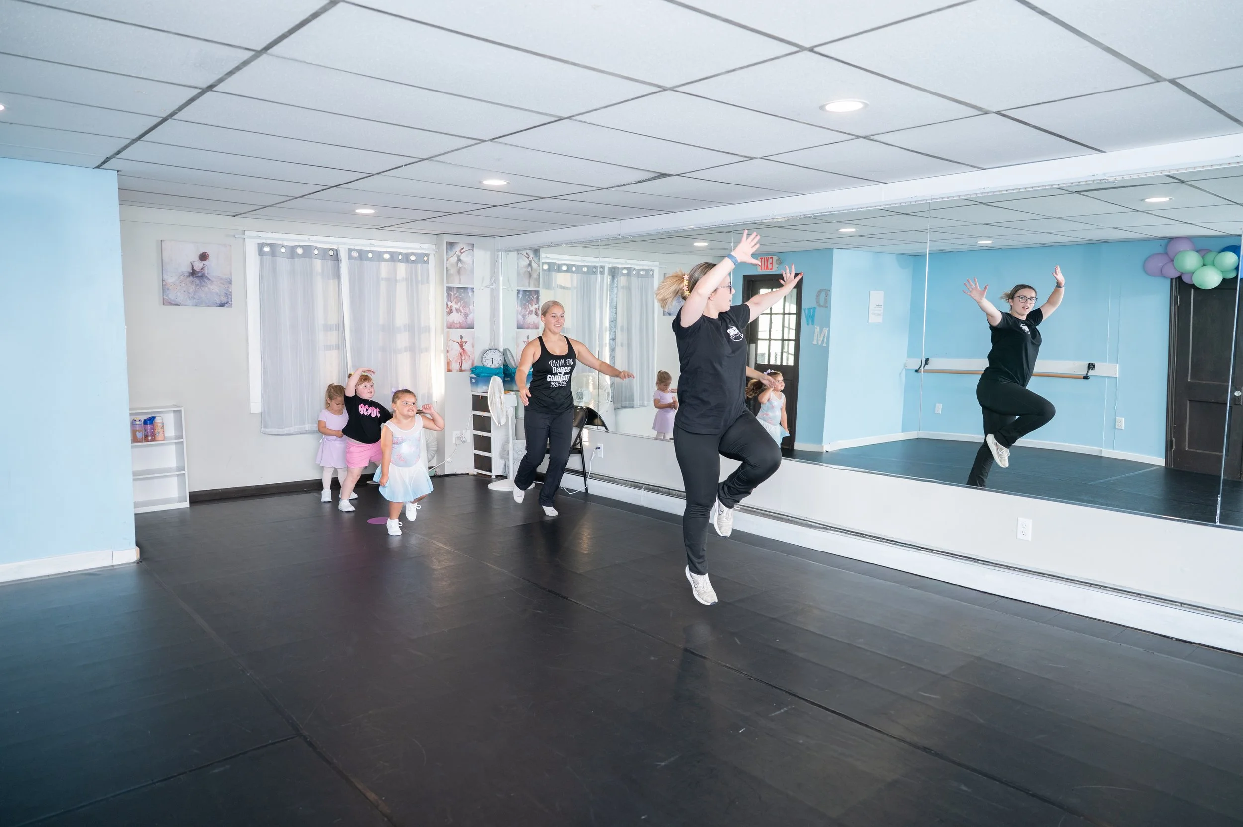 Dance instructor leading young girls in a dance class in a studio with a large mirror, ballet barre, and decorations.