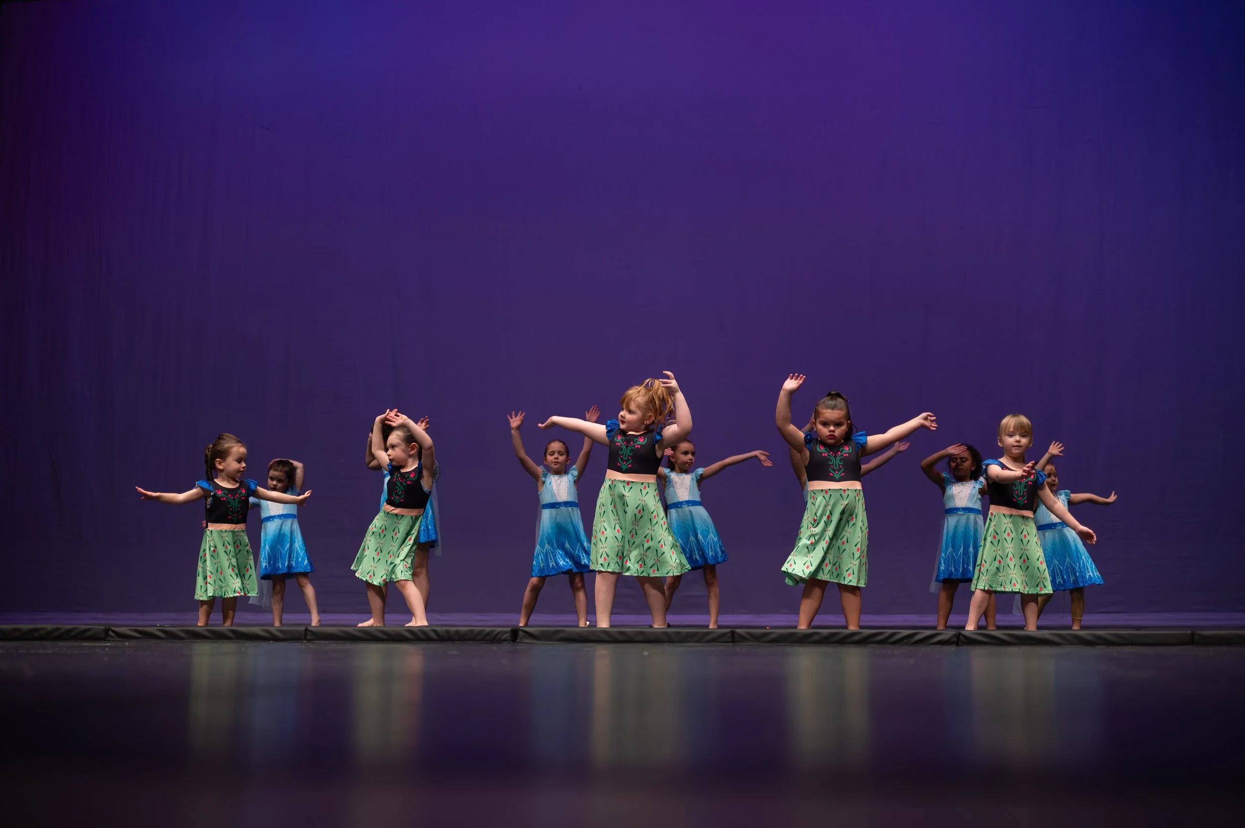 Children’s dance performance on stage with girls wearing colorful dresses, dancing against a purple background.