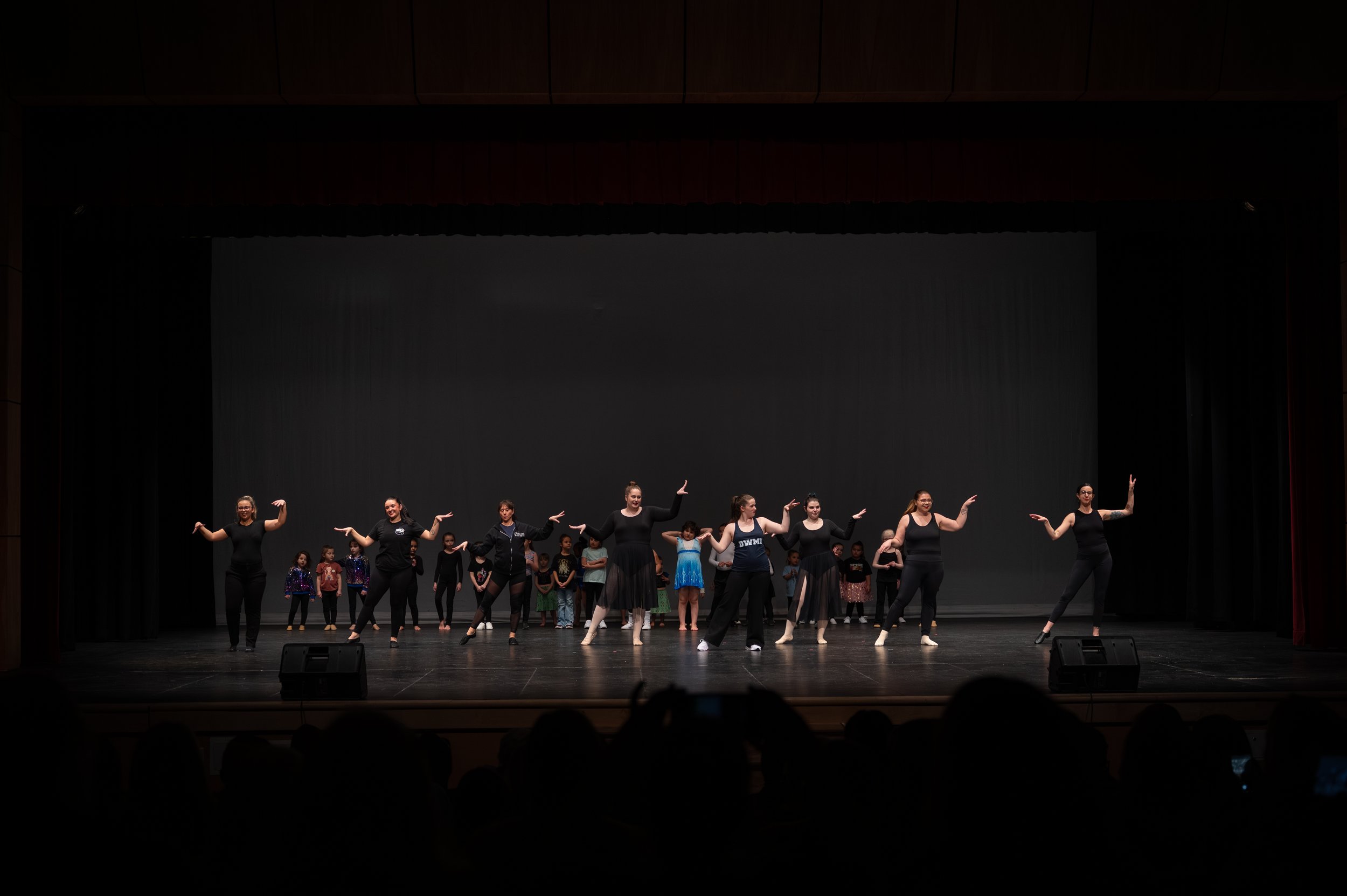 A group of dancers, mostly women, performing a dance on stage with children standing behind them.