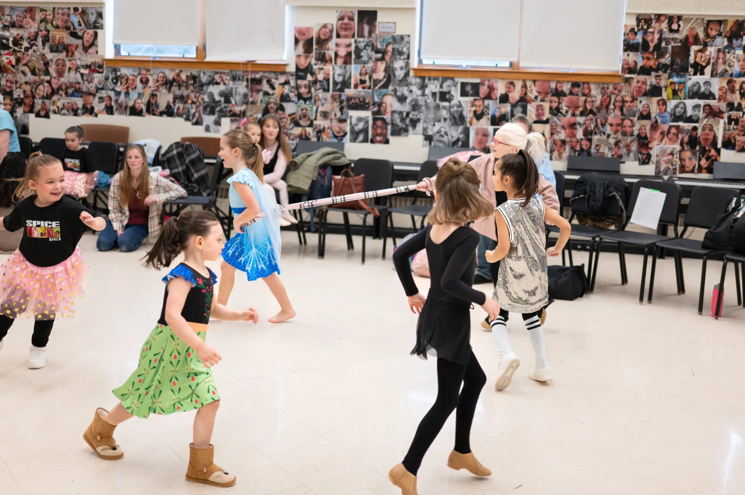 Children dancing in a classroom with wall decorations made of photos of faces and people.