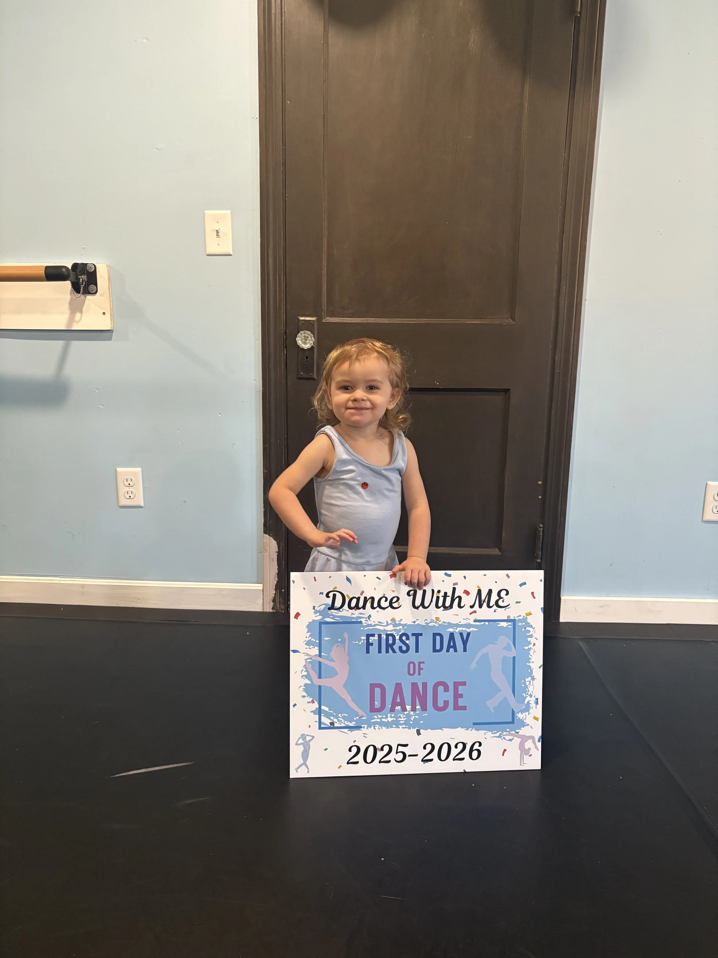 A young girl with curly blonde hair and a blue dance costume, standing indoors and holding a sign that reads 'Dance With ME, First Day of Dance 2025-2026'.