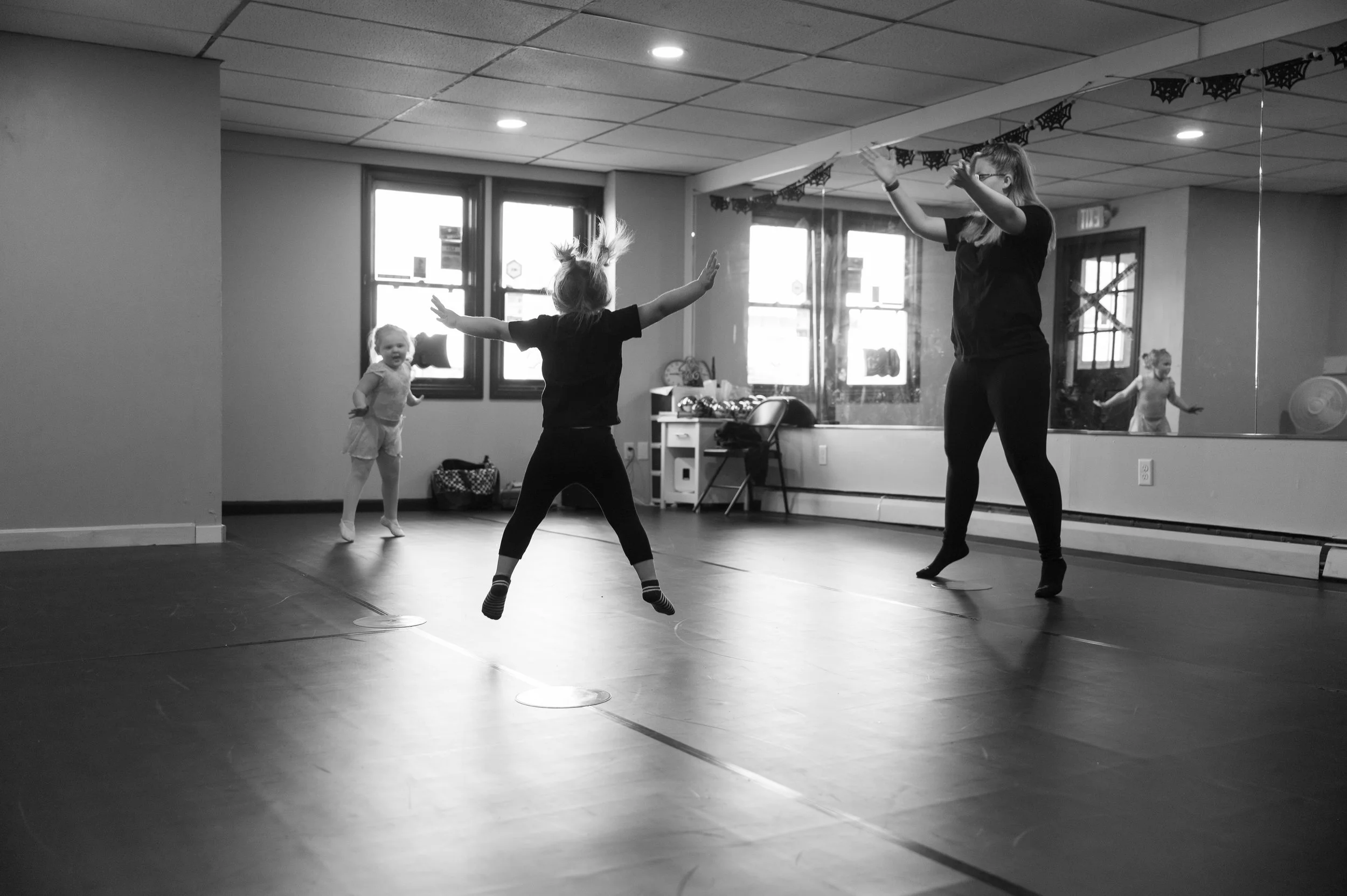 A dance class for young children with an instructor leading two children in a dance studio with mirrored wall and windows. One child is jumping with arms outstretched, and another child is smiling and moving nearby.