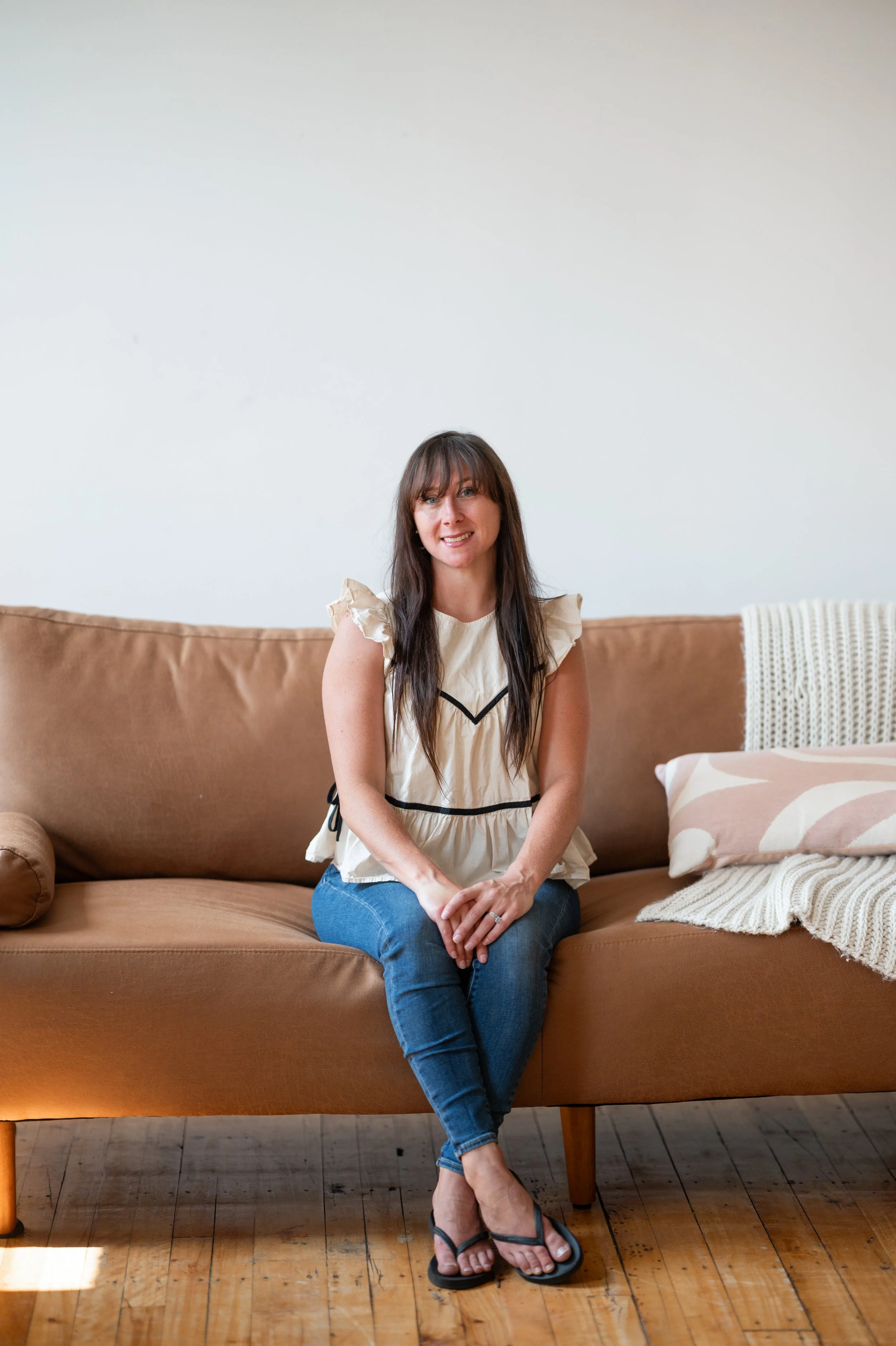 A woman with long dark hair sitting on a tan sofa in a room with wooden floors, wearing a cream top with ruffled sleeves and blue jeans, smiling at the camera.