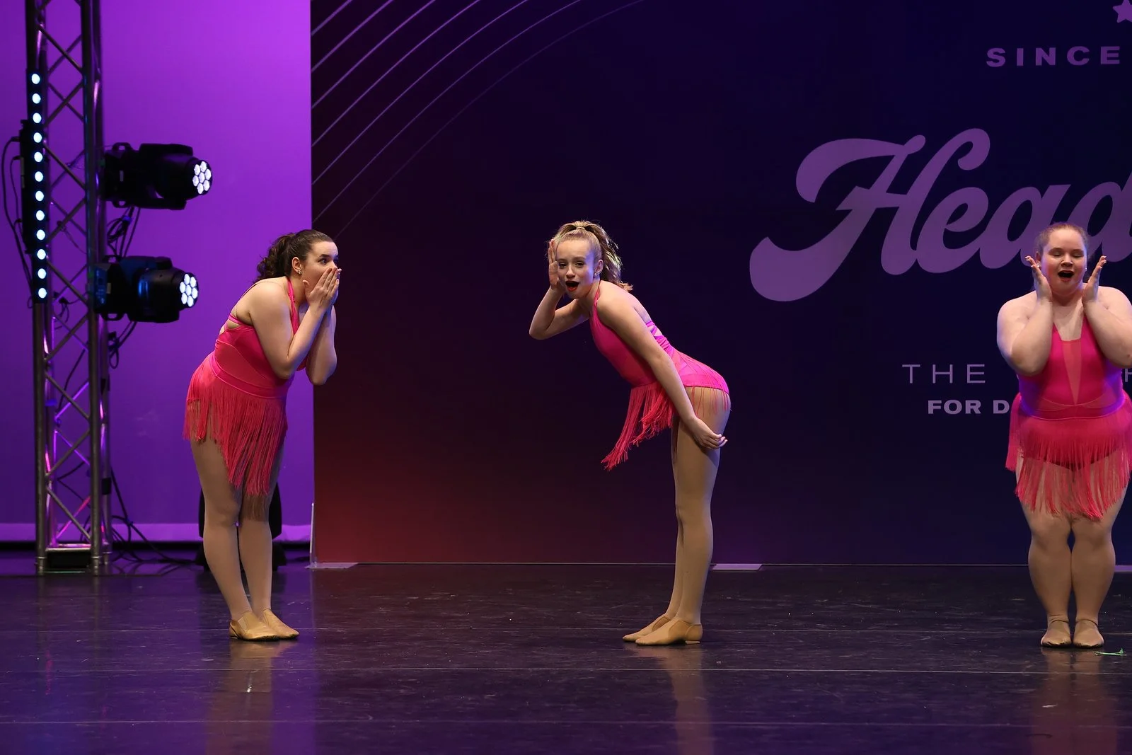 Three young female dancers in pink costumes with fringe, wearing beige dance shoes, stand on a stage with purple and black background. They appear surprised with hand gestures covering their mouths or cheeks, during a performance or competition.