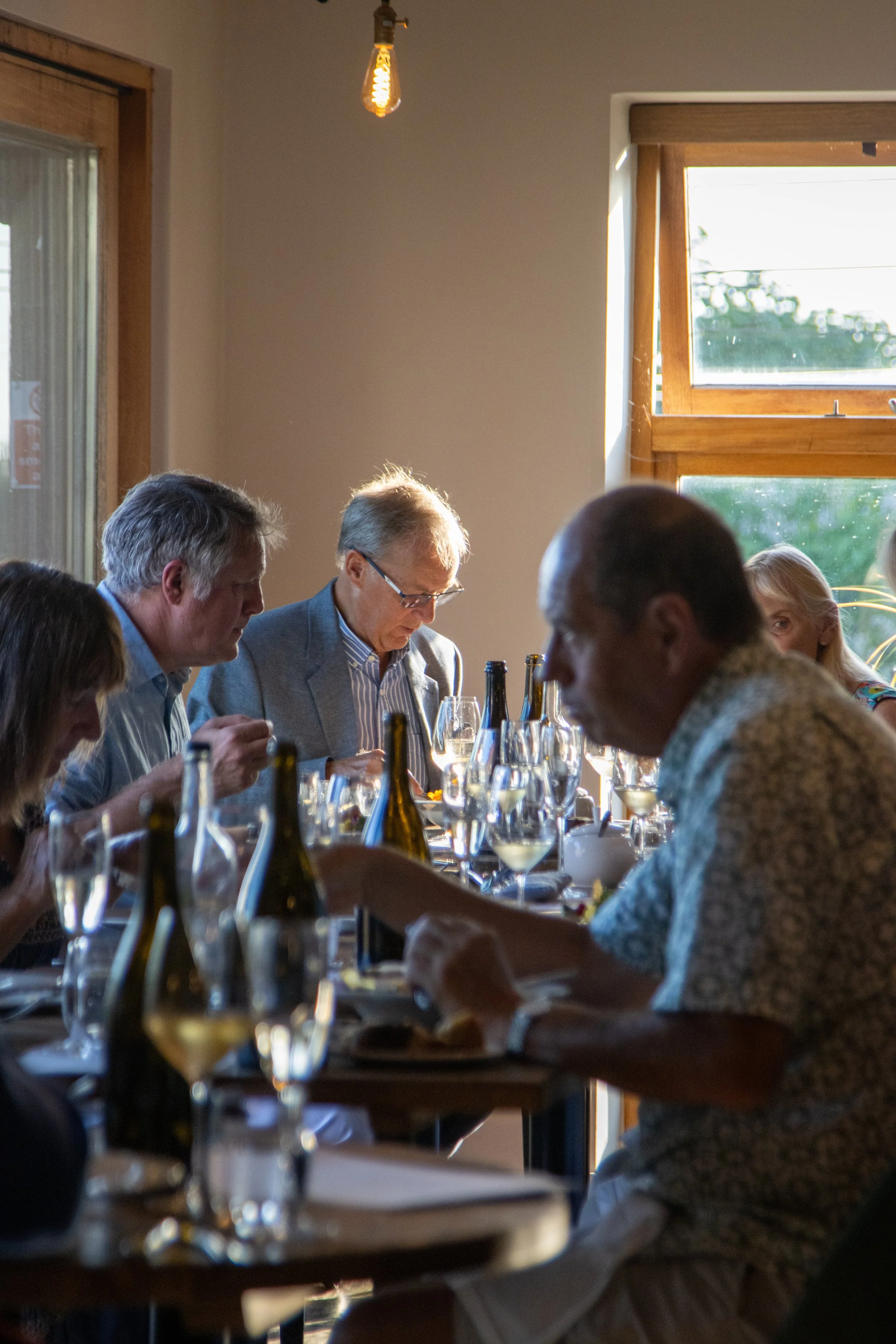 People sitting at a dining table enjoying a meal during daytime, with wine glasses and bottles, in a room with wooden window frames and an exposed light bulb hanging from the ceiling.