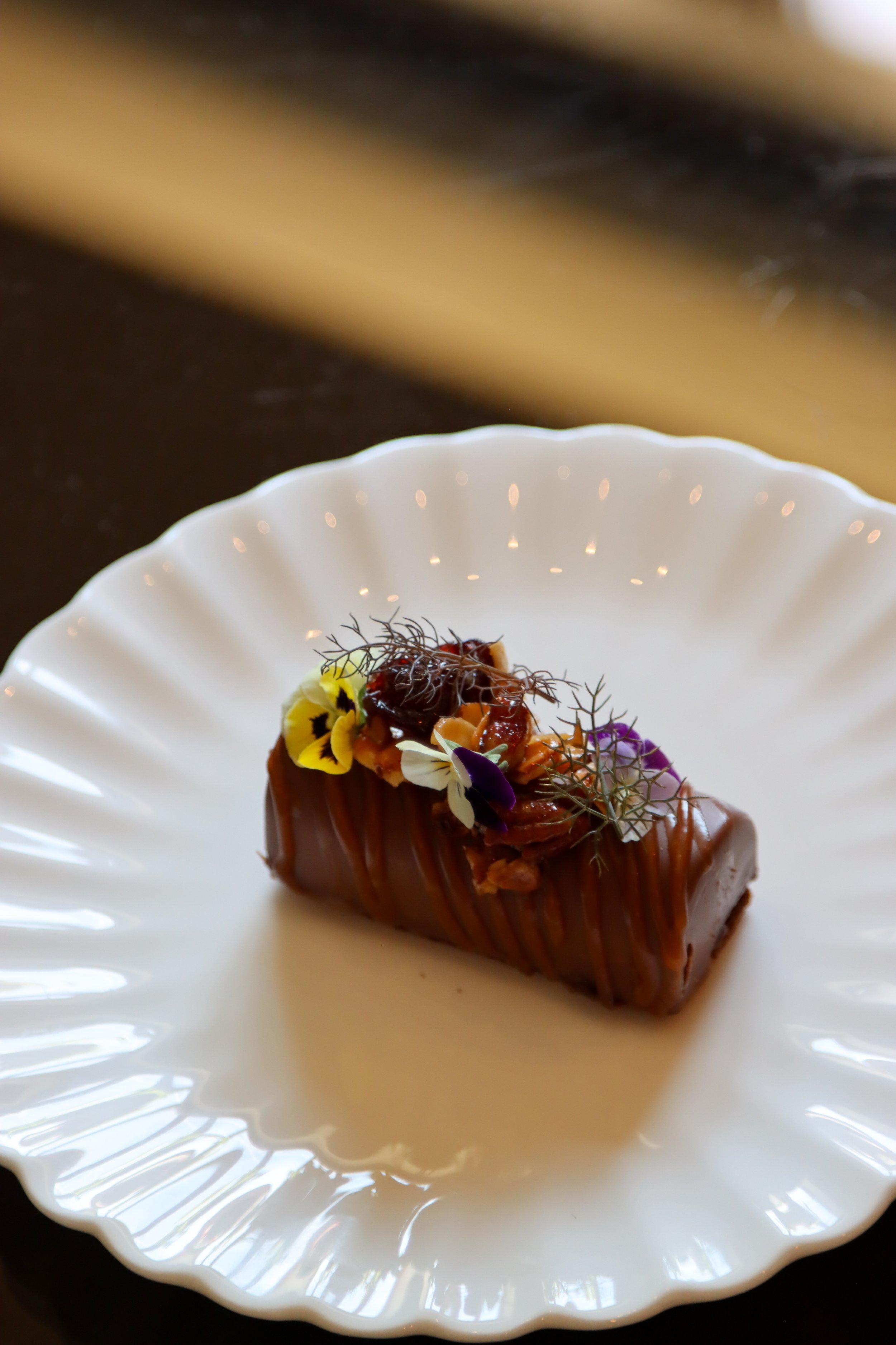 A close-up of a plated dessert featuring a cube of glazed chocolate cake, decorated with edible flowers and microgreens on a white, scalloped-edge plate.