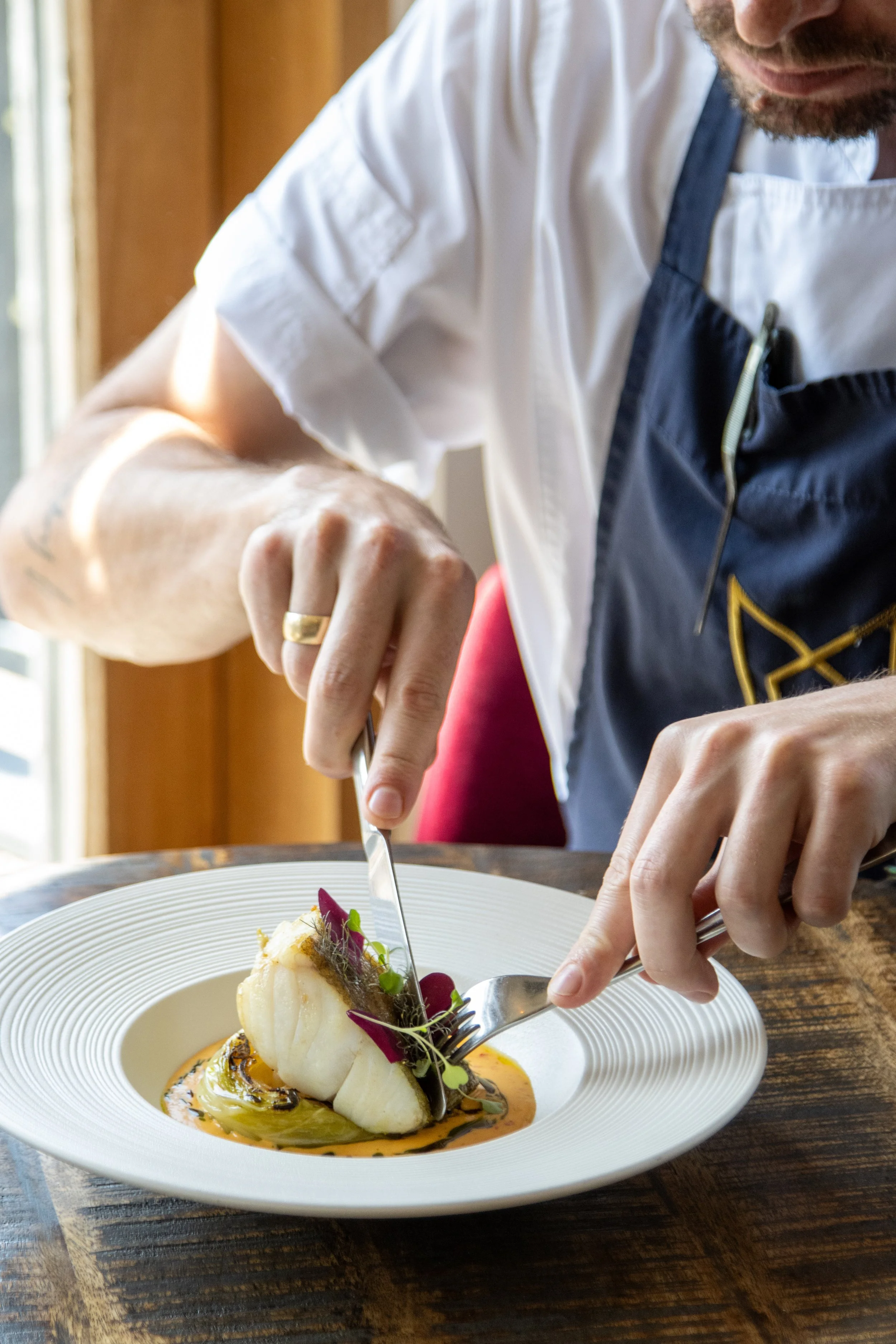 Close-up of a chef in a white uniform and apron garnishing a plated gourmet dish with herbs and edible flowers on a white plate, set on a wooden table.