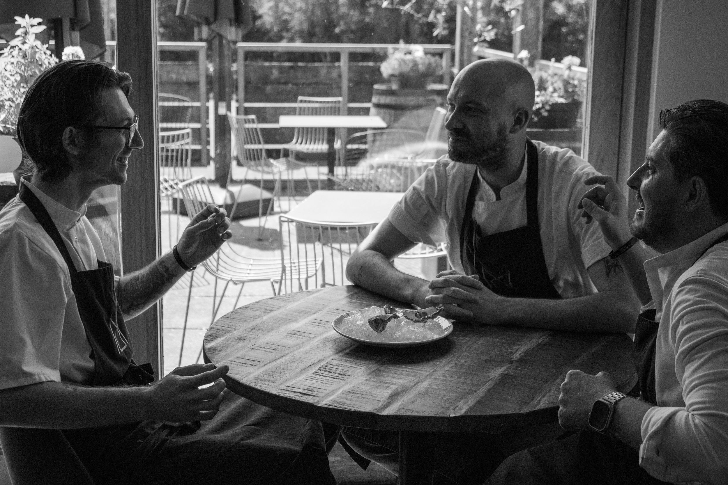 Four people of diverse backgrounds sitting at a round wooden table in a restaurant, engaged in conversation. A plate with food is in the center. Large windows behind them reveal an outdoor patio with chairs, tables, plants, and a wooden fence. The scene is in black-and-white.
