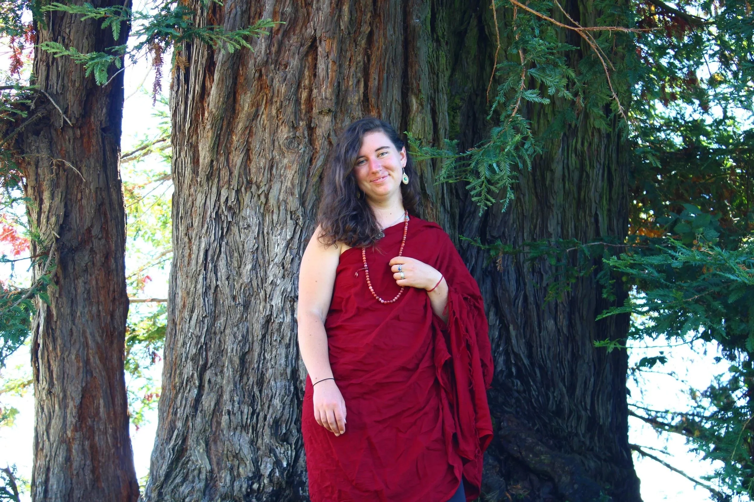 A woman with long curly hair, wearing a red dress and jewelry, standing outdoors in front of large tree trunks and green foliage.