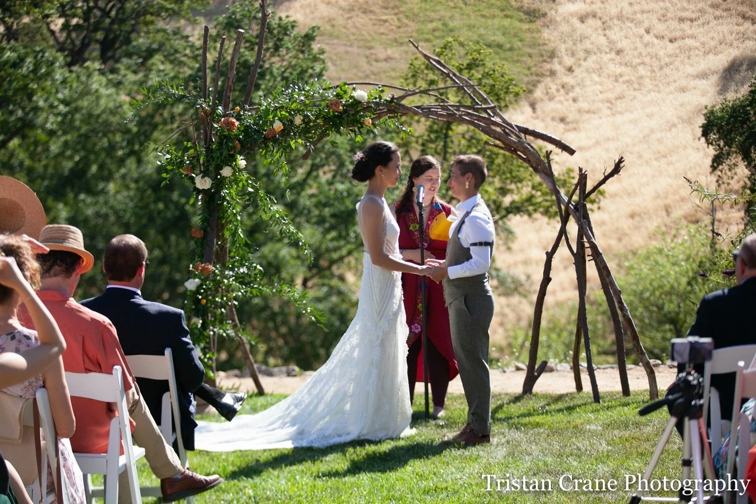 A couple getting married outdoors under a rustic archway made of branches and flowers. The bride is in a white gown and the groom in a gray suit, holding hands and facing each other. Guests seated on white chairs are watching the ceremony, and a photographer is nearby. The setting is on a grassy area with trees and a hillside in the background.