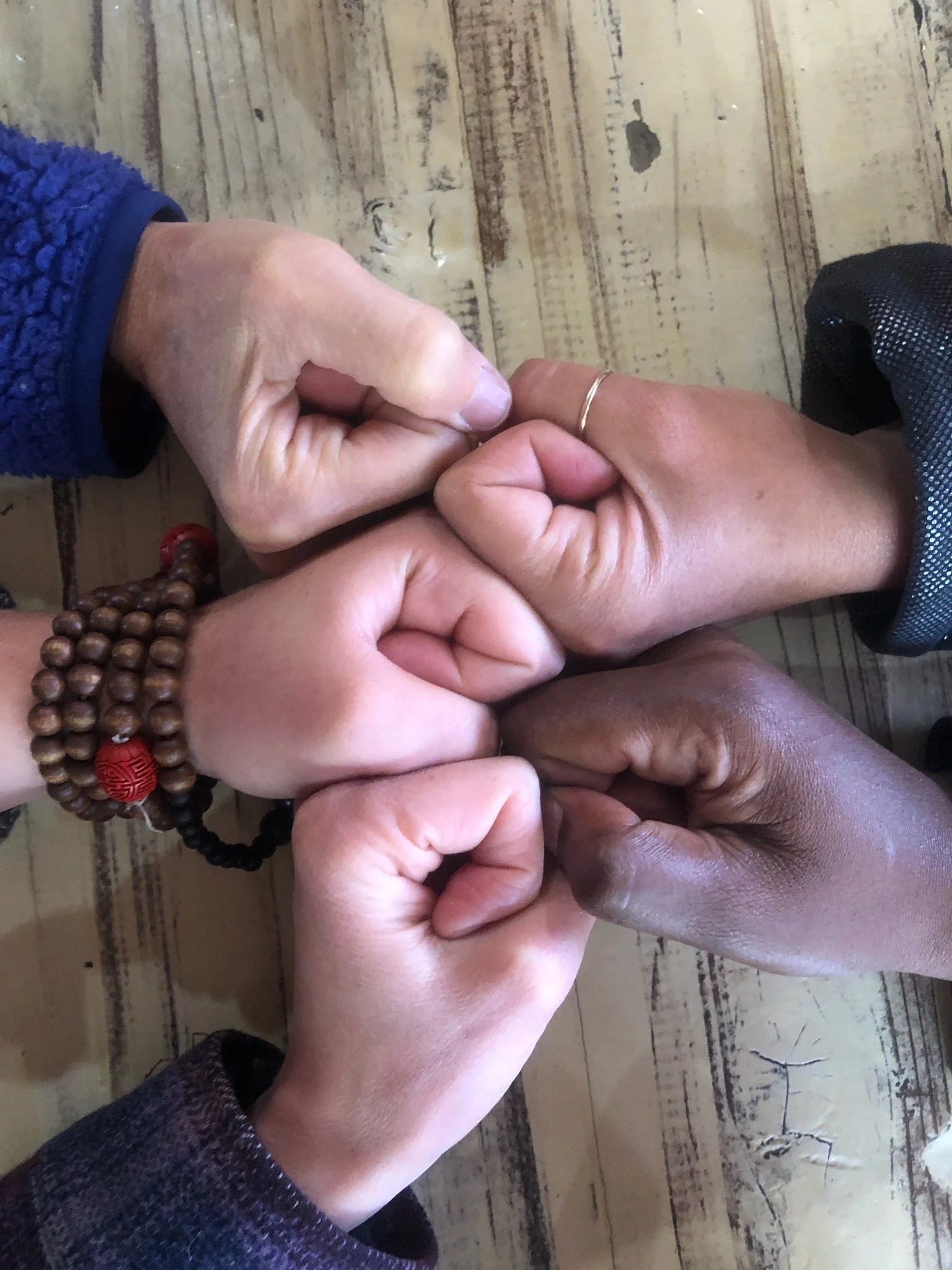 Multiple hands of different skin tones holding each other's fingers, symbolizing connection or support, with a wooden floor background.