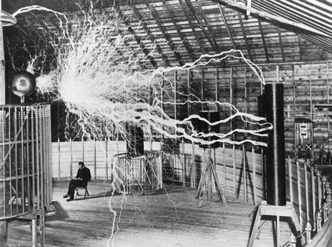 Tesla coil generating electrical sparks inside a wooden building with a seated man observing.