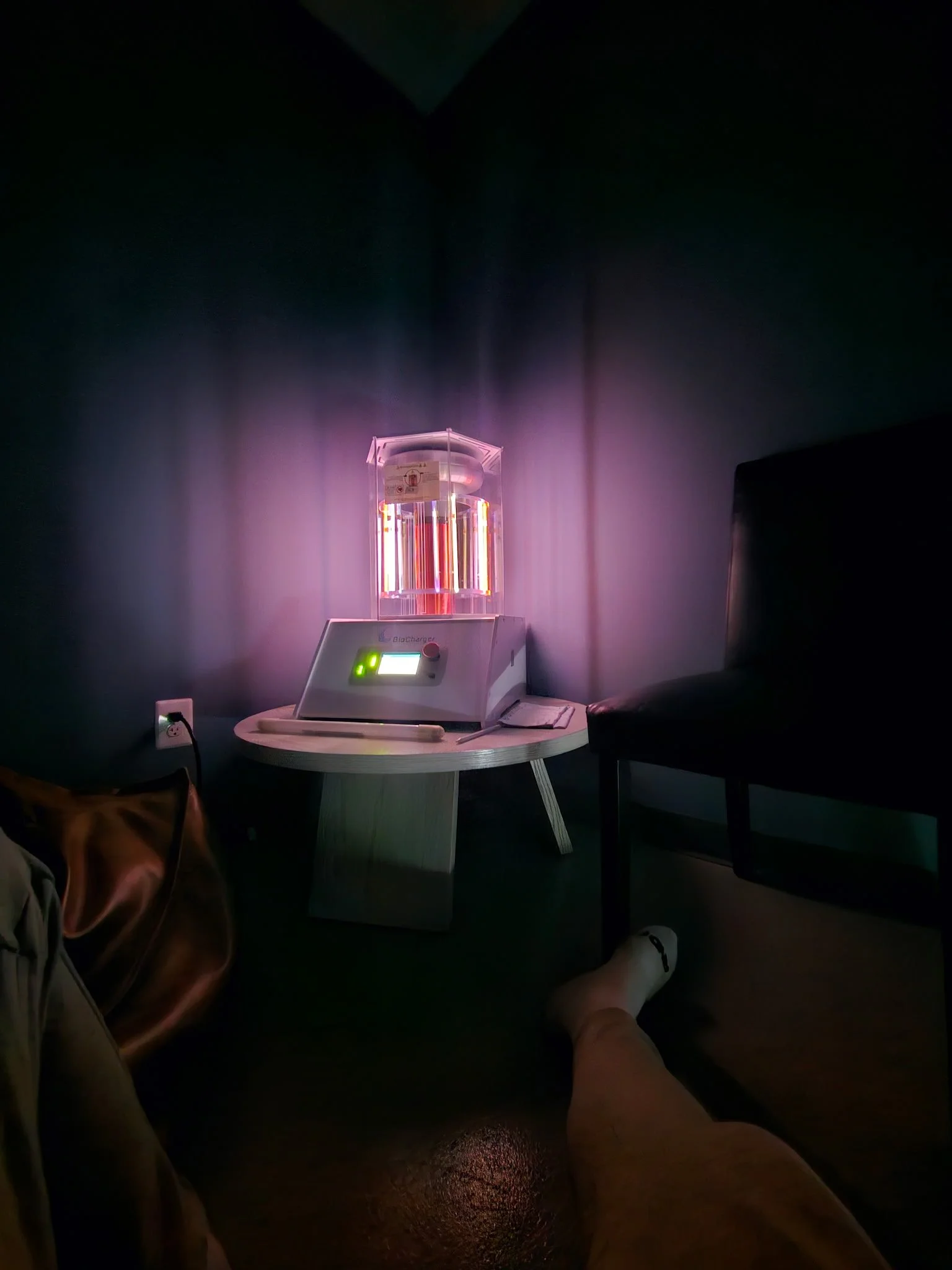 A person lying on a chair or bed, with their legs visible, is near a small round table. The table holds a UV light sanitizer device turned on, emitting purple and pink light. The room has dark walls and a dark floor, with an electrical outlet visible on the wall.