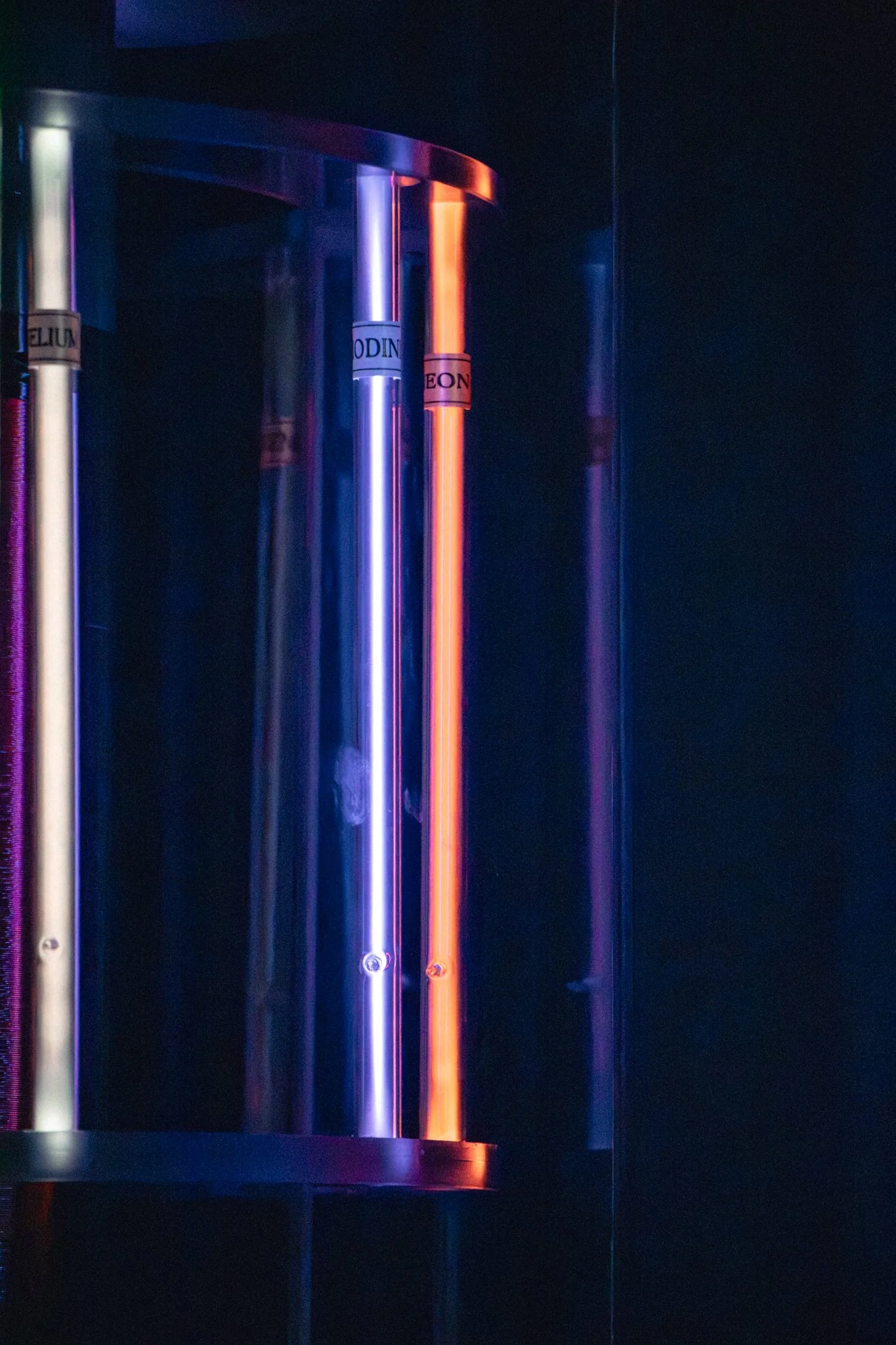 Close-up of three glowing tubes, one yellow, one purple, and one orange, mounted on a dark wall with a blue and faint purple background.