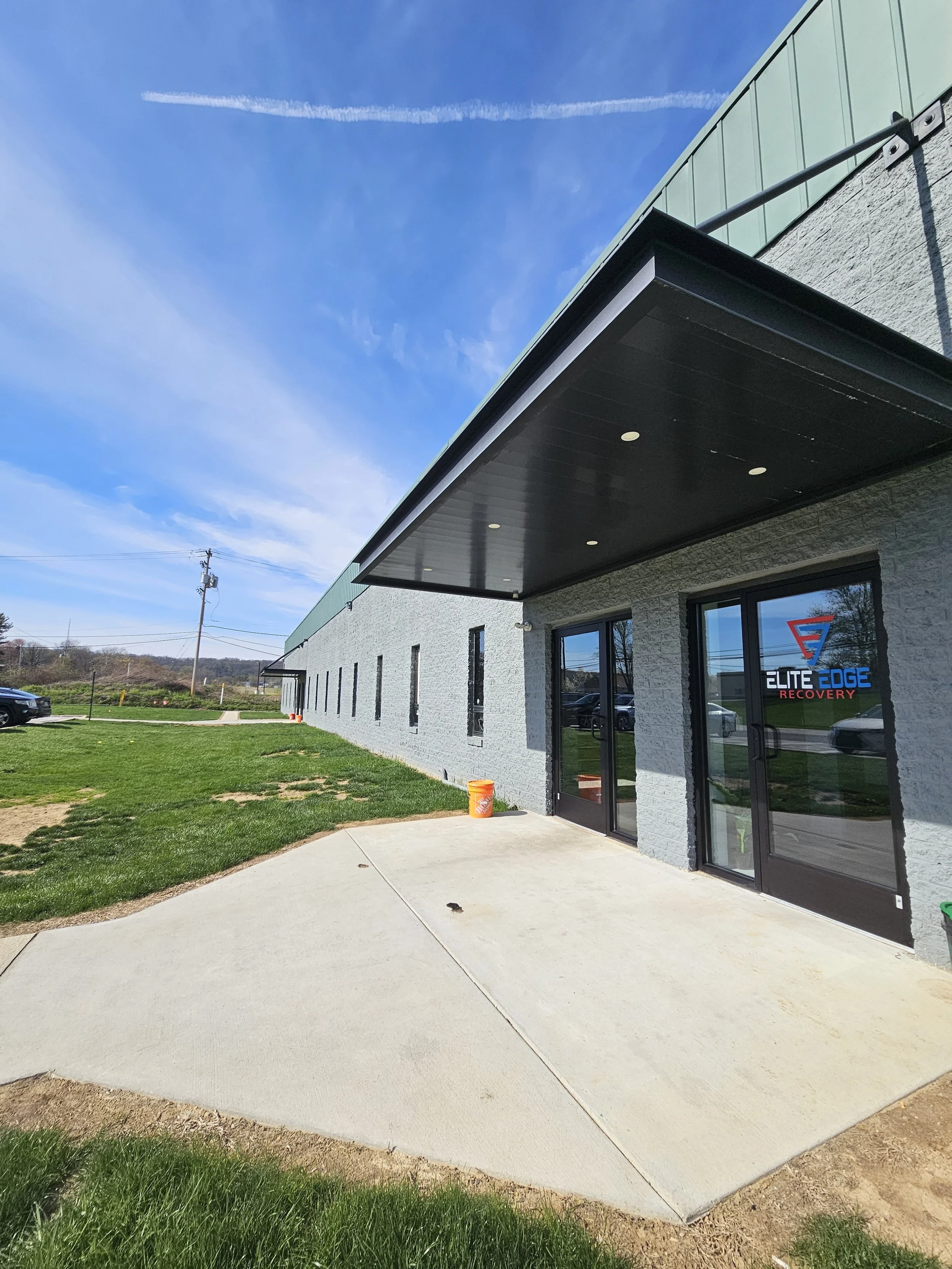 Exterior of a commercial building with a concrete sidewalk, grass, and a sign that reads 'Elite Edge Recovery' on the window, under a blue sky with some clouds.