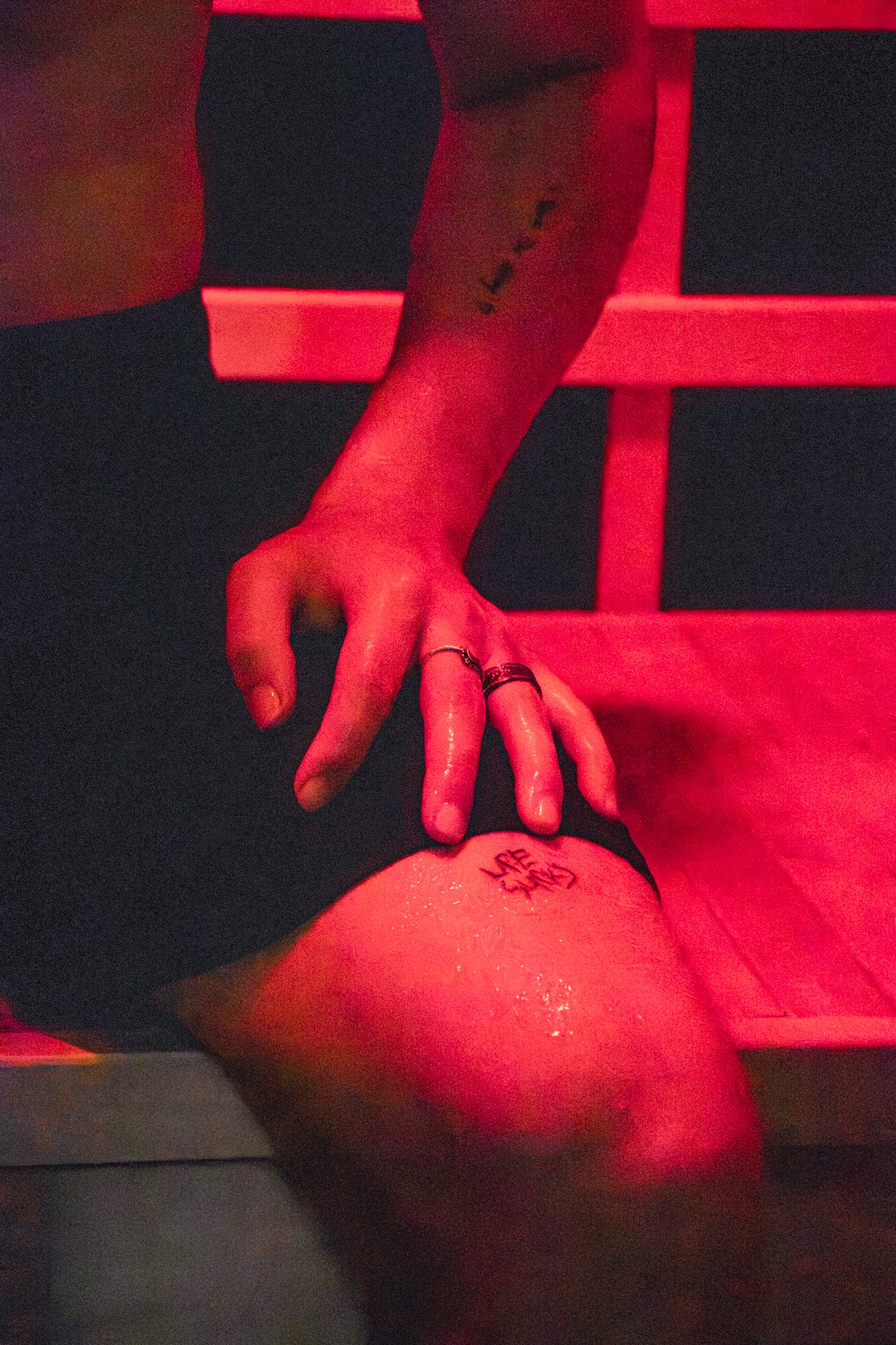 A person's hand with rings and tattoos reaching towards their knee, which has a tattoo of a flower. The scene is illuminated with red lighting, and the background shows a red chair or bench. the person is in an infrared sauna.