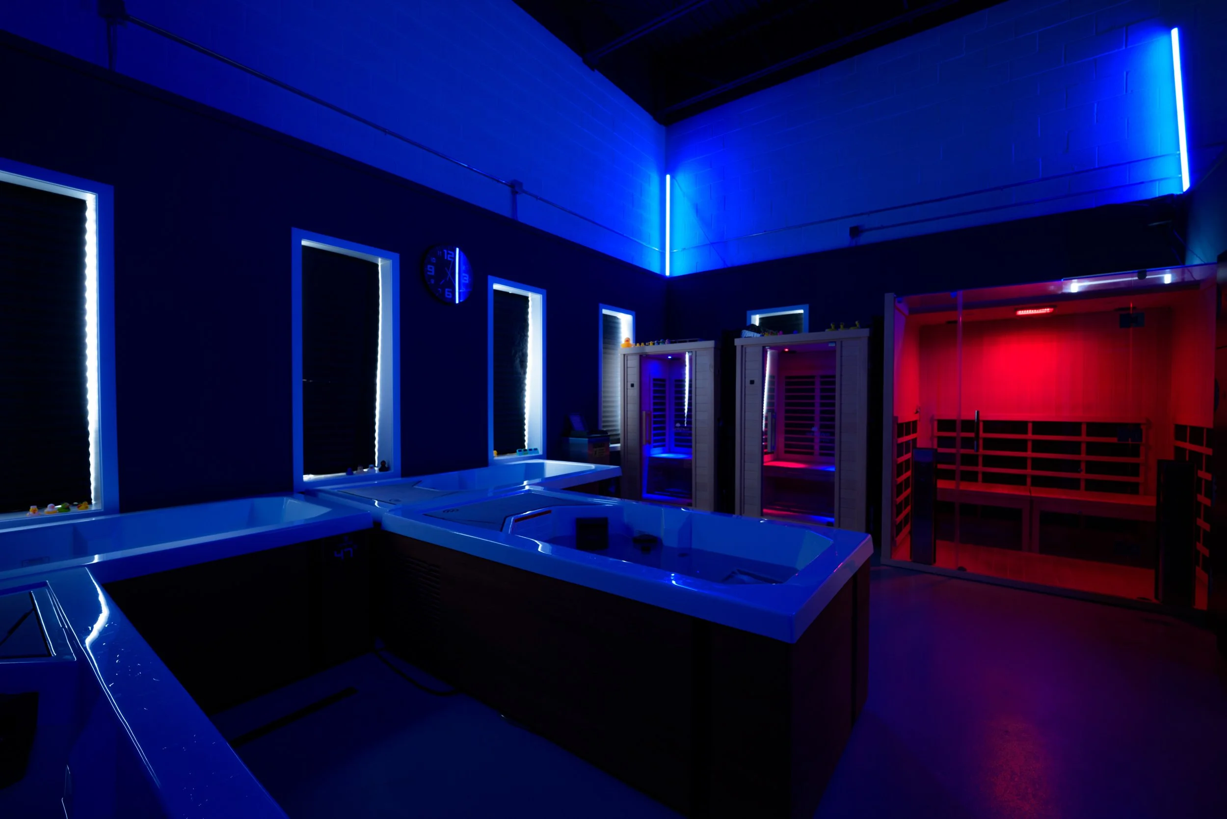 A modern spa room illuminated with blue and red LED lights, featuring hot tubs, sauna cabins, and a wall clock.