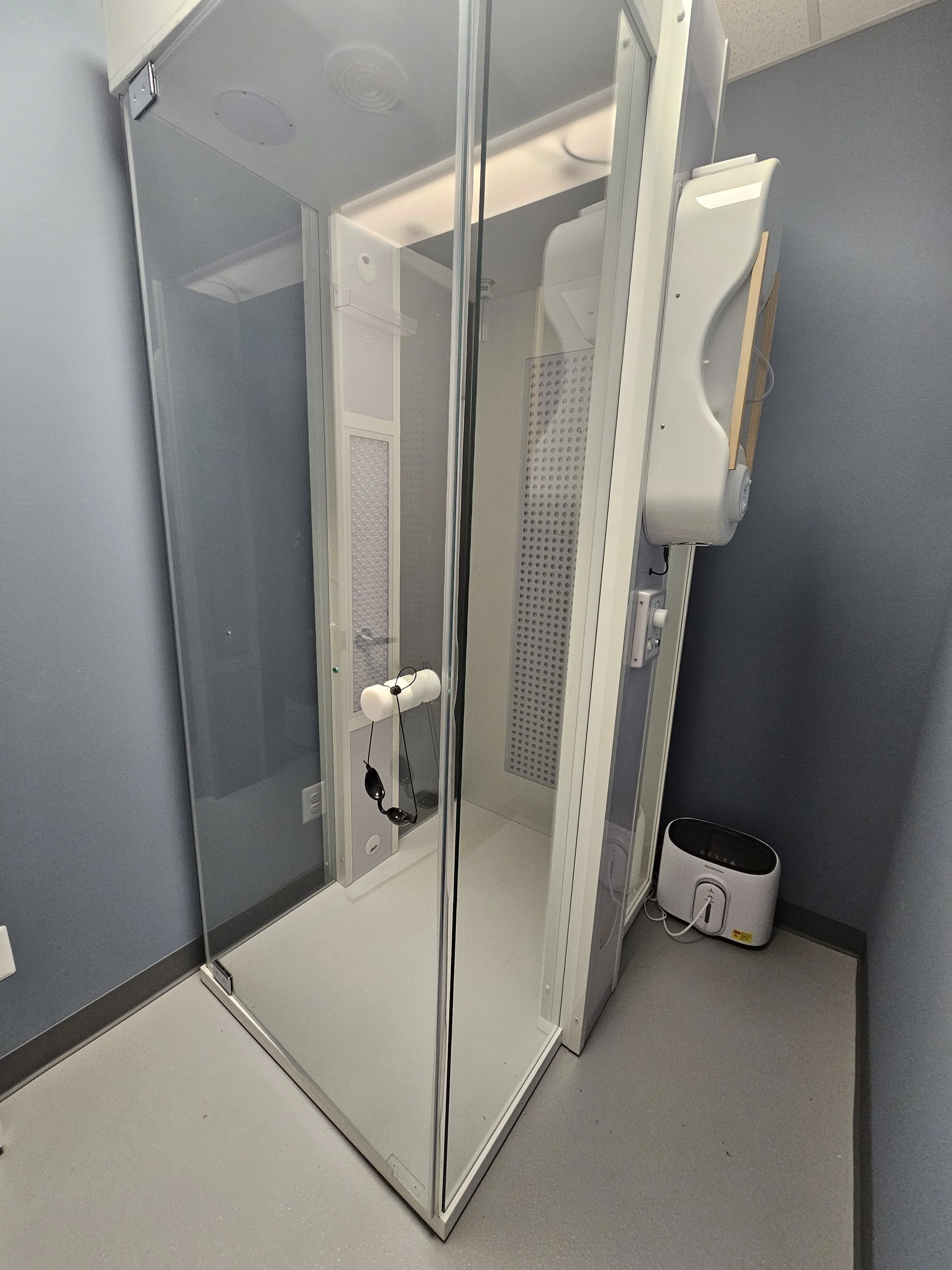 A modern medical shower cabin with glass doors, attached to a wall with controls and a soap dispenser, in a clinical setting.