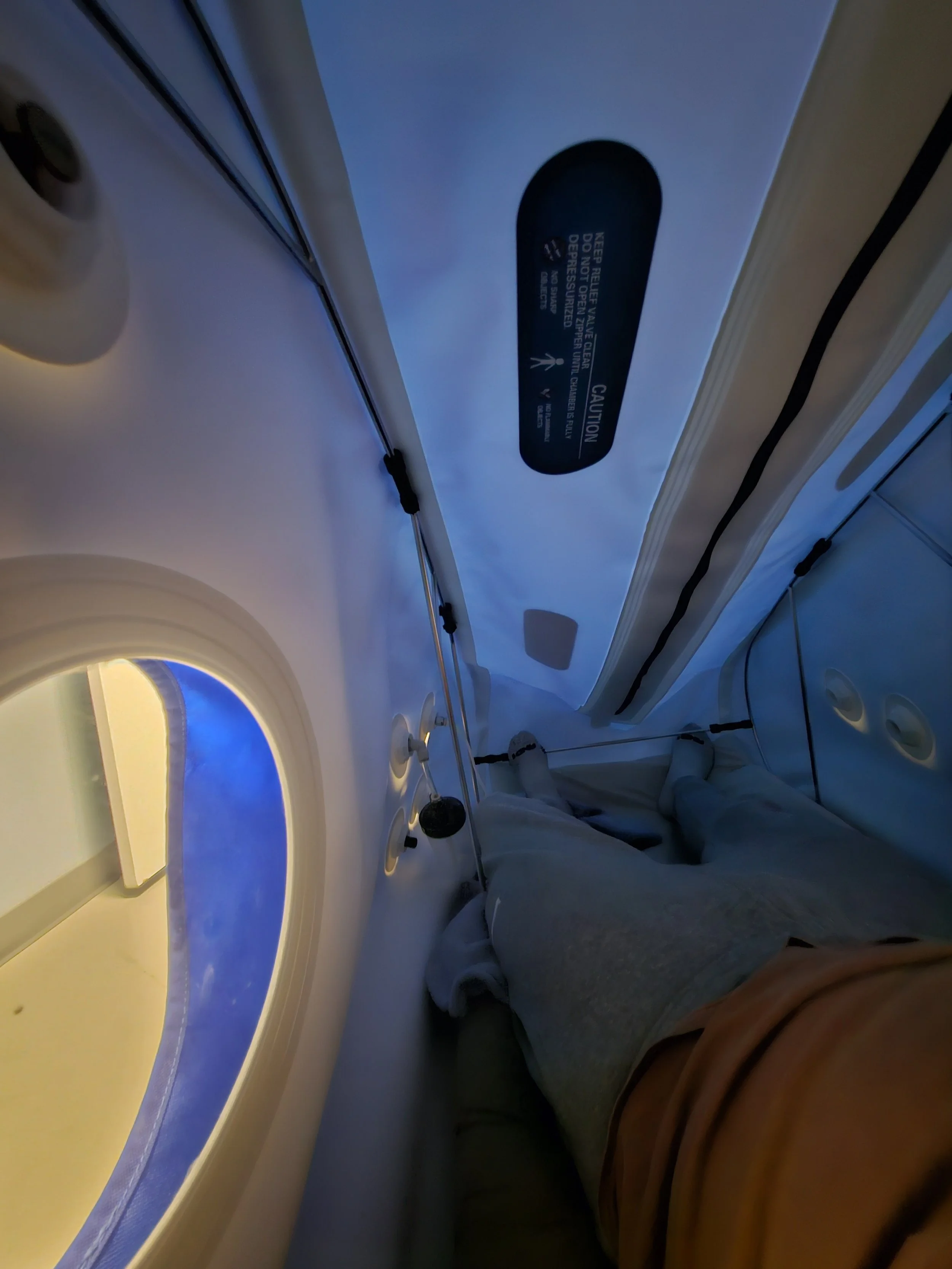 Photo taken inside a hyperbaric oxygen chamber showing a person's legs and feet with gray sweatpants and orange shorts, with a window to the left and a safety instruction label on the ceiling.