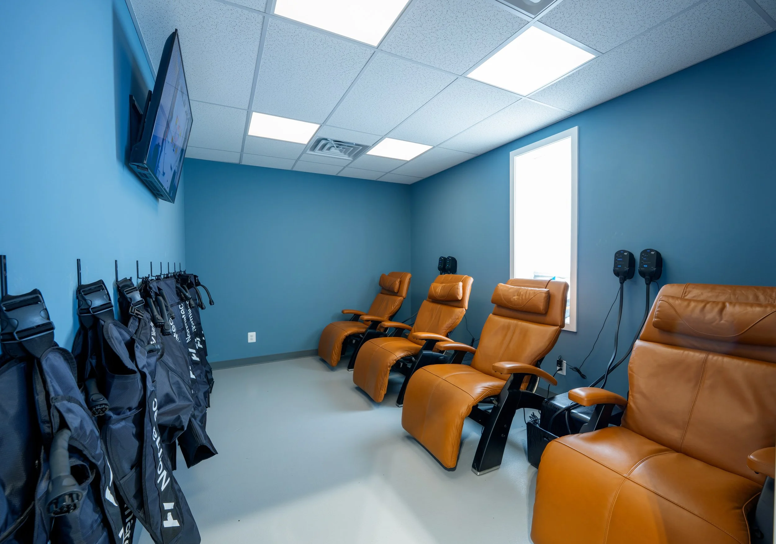 A meditation or relaxation room with four brown leather reclining chairs, two infrared therapy devices on the wall, a row of black golf bags on hooks, and a wall-mounted television on a blue wall.