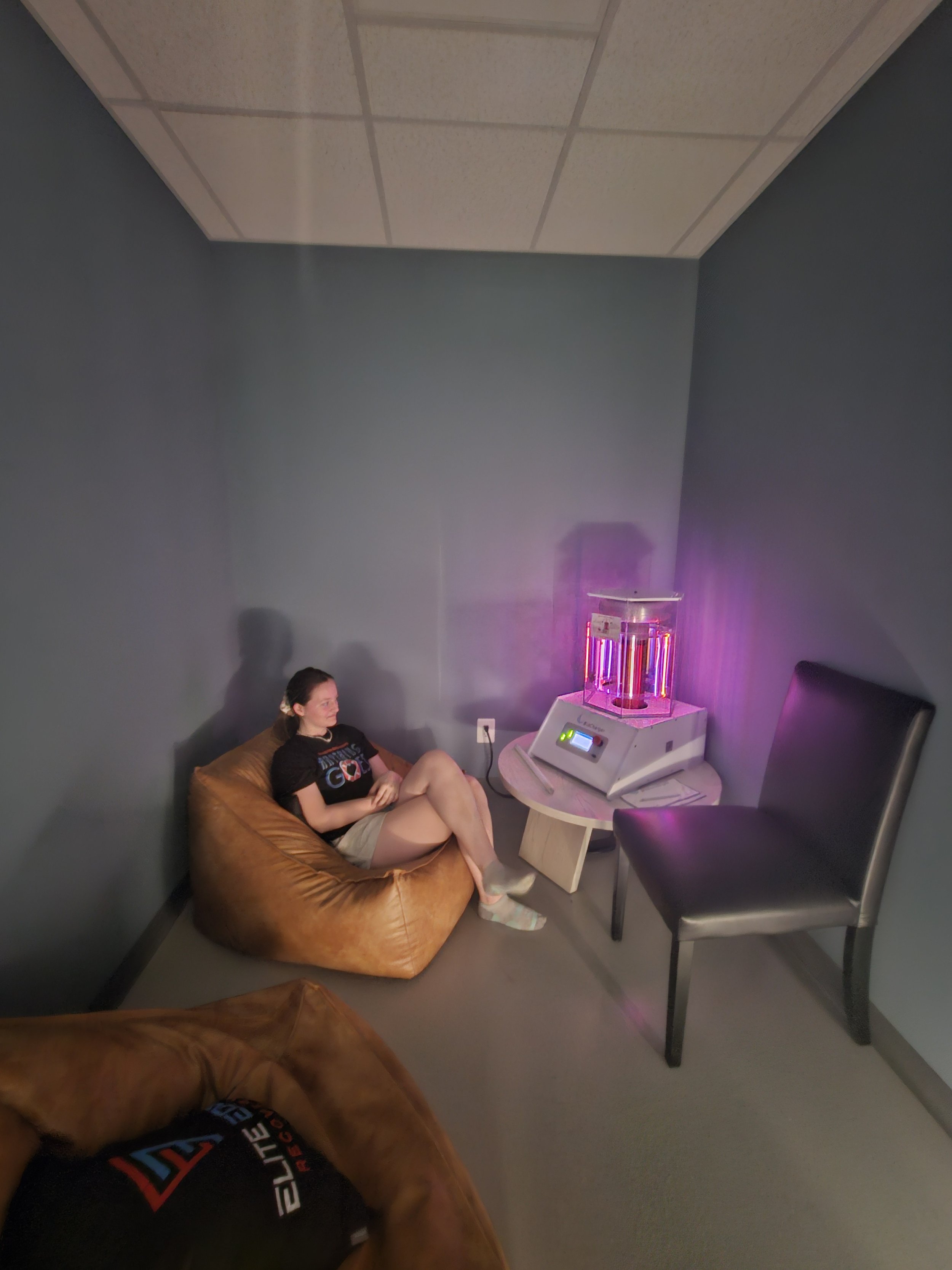 A woman sitting on a brown bean bag chair in a small room, looking at a pink and purple ultraviolet light therapy device on a small table. There is a second black chair in the corner and a person's arm visible in the foreground.