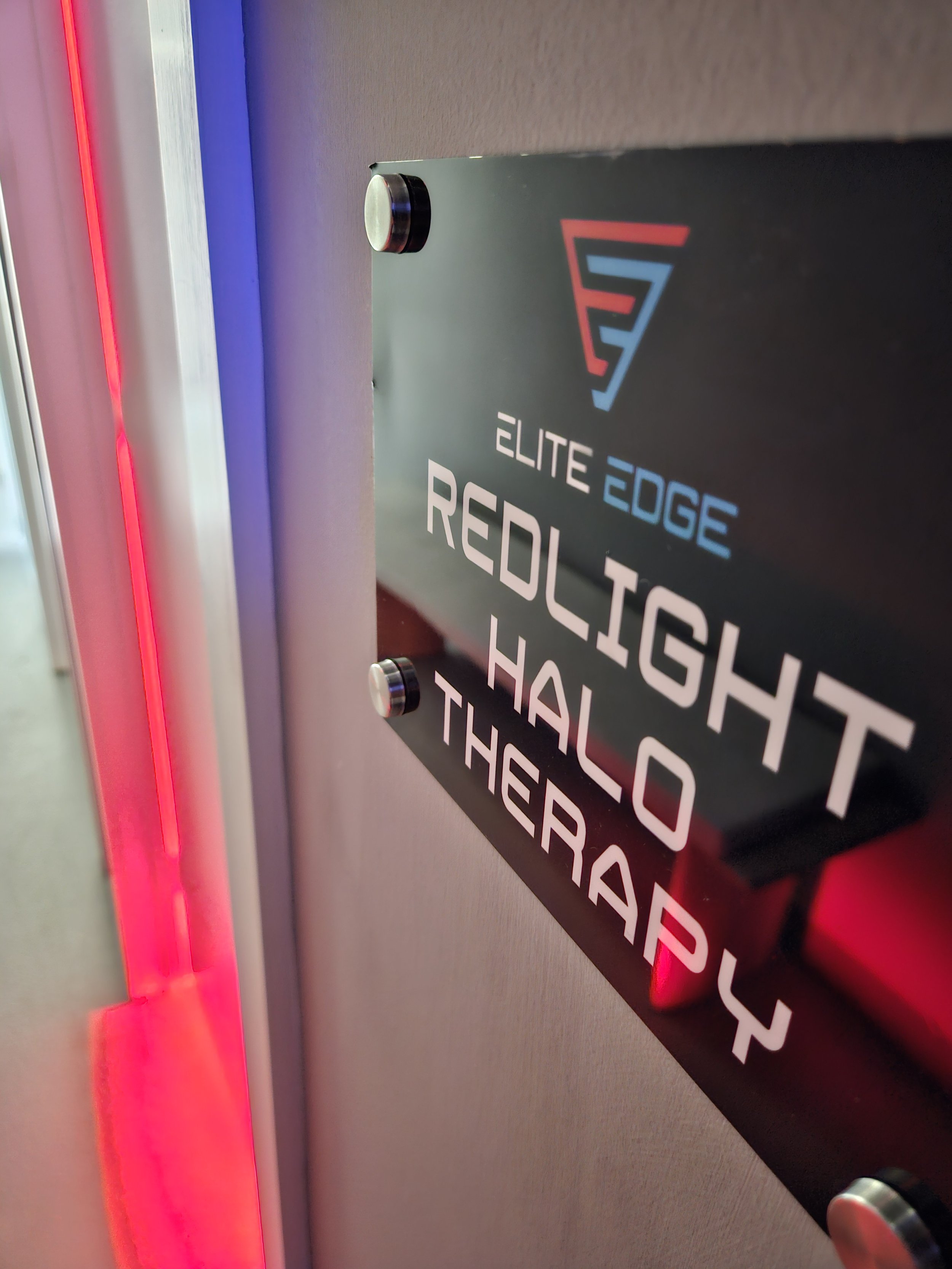 Close-up of a sign on a wall that reads 'Elite Edge Red Light HALO Therapy' with a logo at the top. The sign is mounted next to a wall with red and blue lighting accents.