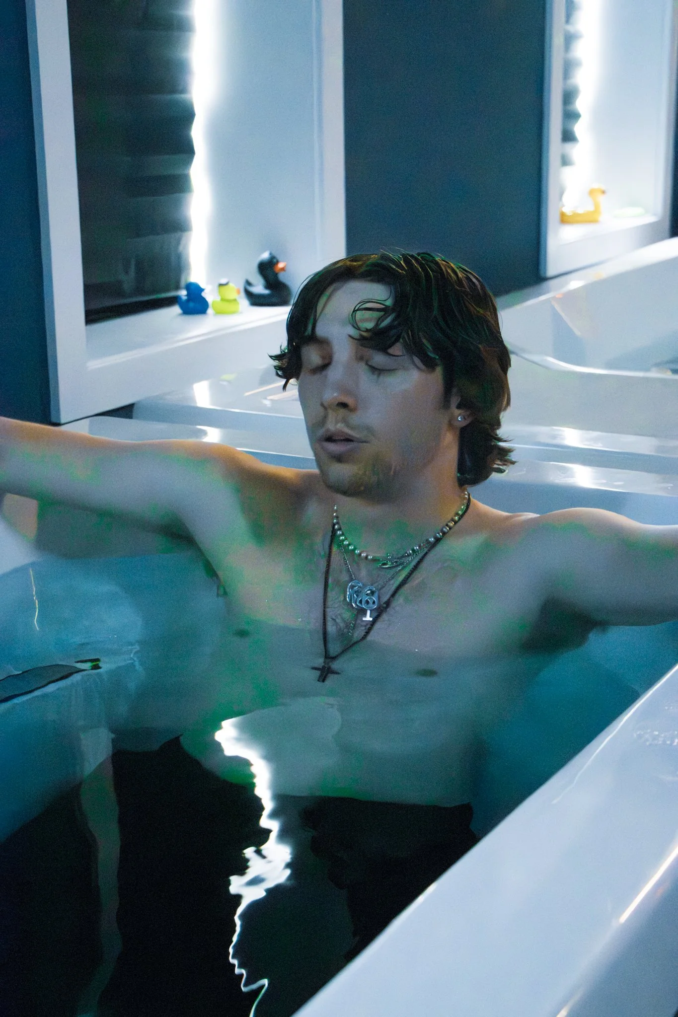 A young man with dark, wet hair relaxing in a Cold Plunge, with his eyes closed and an expression of calmness. The background features a blue wall, and small rubber duck figurines are visible on a shelf.