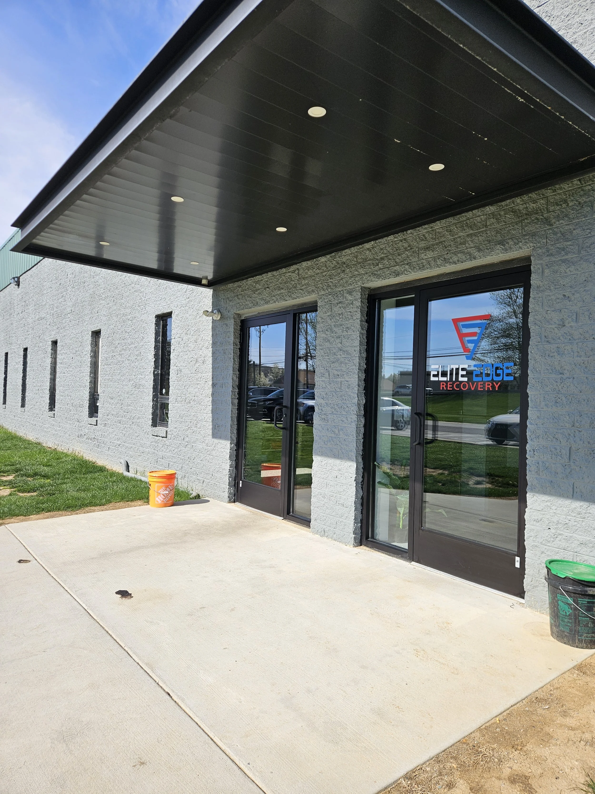 Exterior of a building with gray brick walls, glass double doors with the logo 'Elite Edge Recovery', and a covered entrance with recessed lighting. There are black trash bins, a green bucket, and a small orange bucket outside.