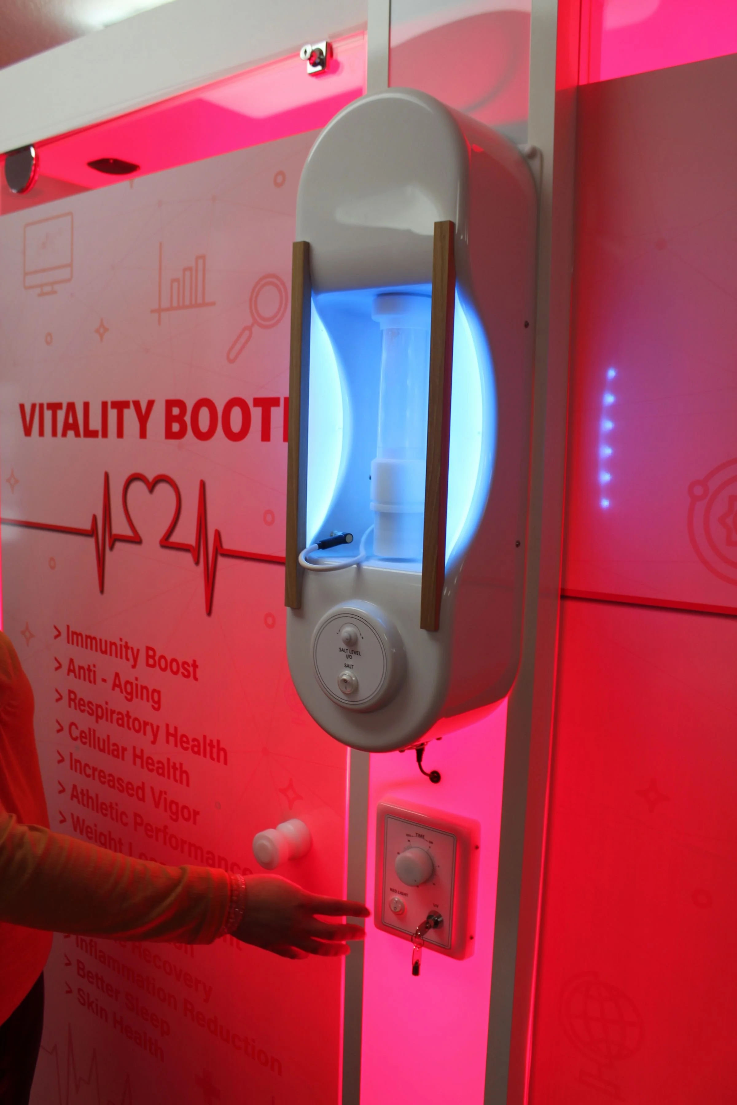 A machine that emits a white and blue light, labeled 'VITALITY BOOTH,' with a leaf salt level indicator and a control panel. The booth is part of a wellness exhibit for a Halo Therapy booth.