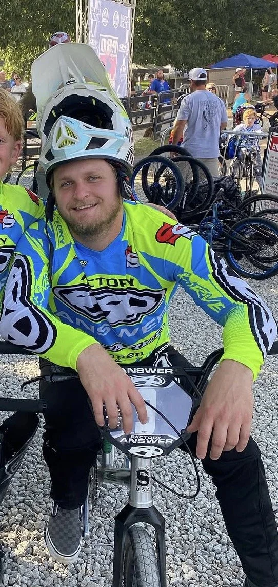 A man wearing a bright cycling jersey and black pants sitting on a bicycle at an outdoor event, holding the handlebars with a helmet on his head, surrounded by other bicycles and people.