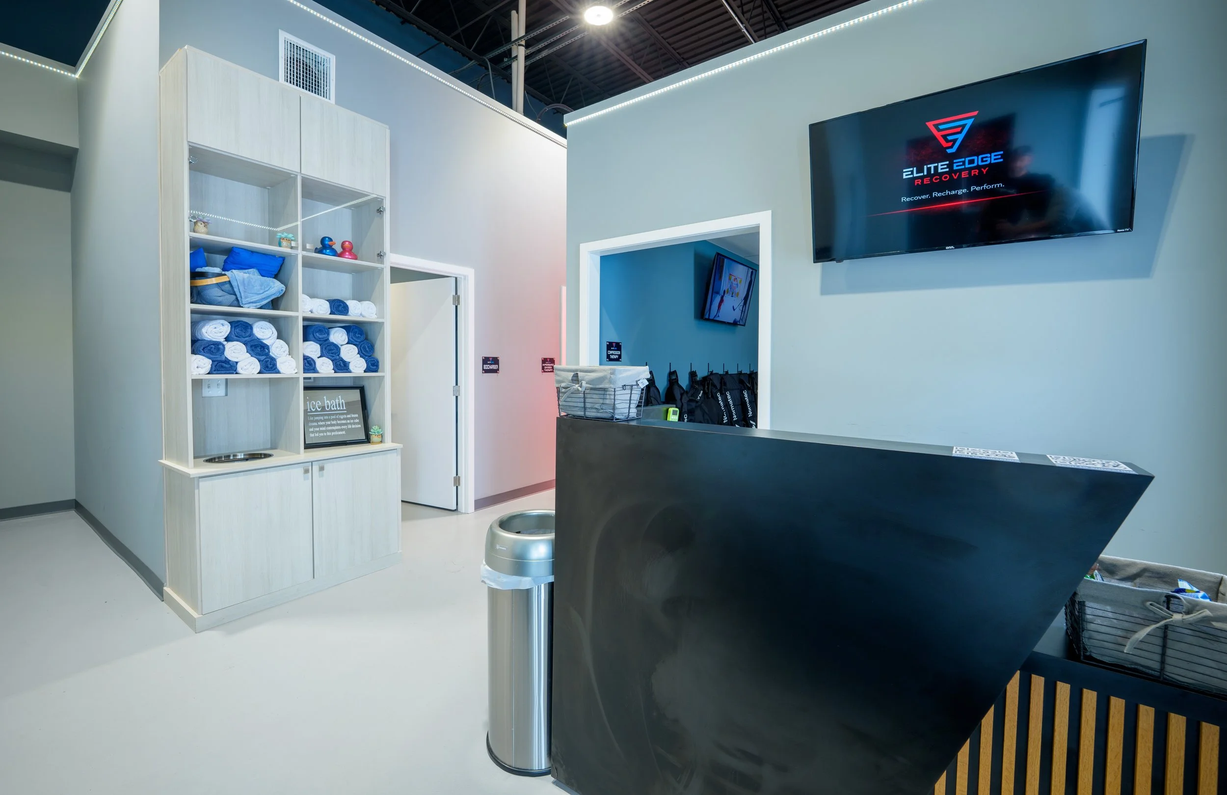 A modern fitness recovery center reception area with a black curved reception desk, a wall-mounted TV displaying 'Elite Edge Recovery', a smaller TV in an adjacent room, a metal trash bin, a shelving unit with rolled towels, and a sign for ice bath services.