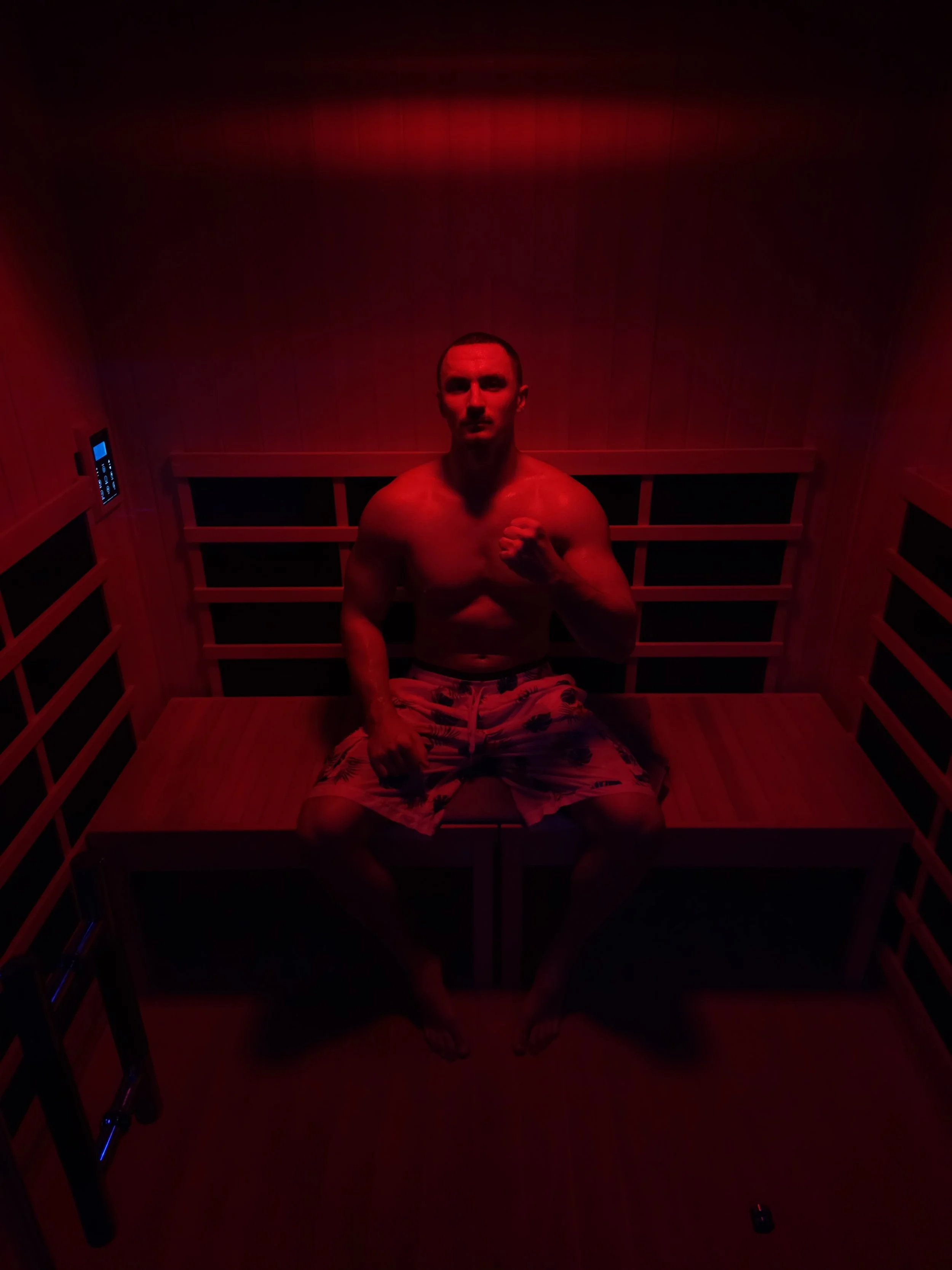 A man sitting shirtless in a wooden sauna illuminated with red lighting, wearing patterned shorts, and making a fist with his right hand.