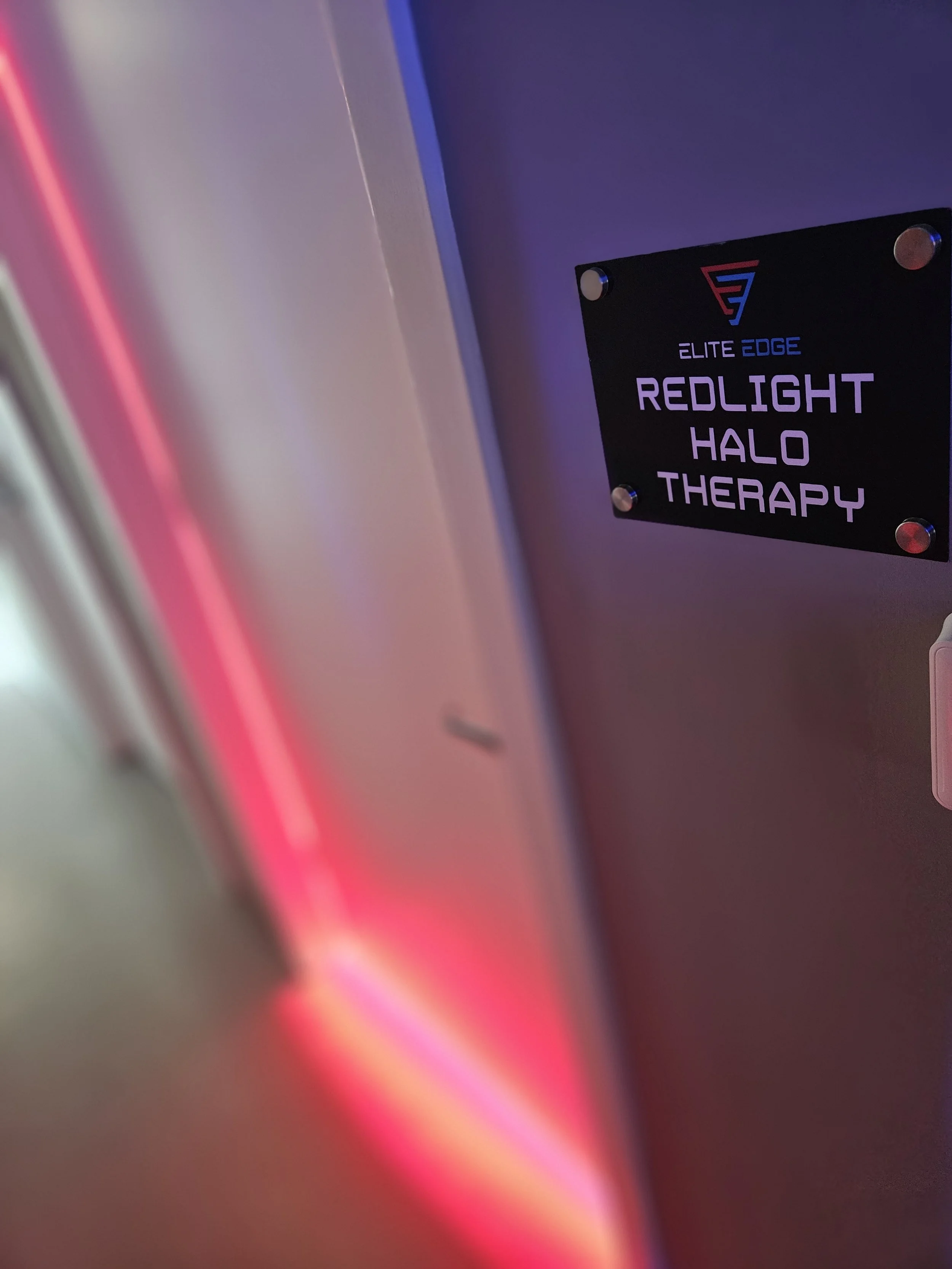 Close-up of a black sign with the logo and text 'Elite Edge Red Light Halo Therapy' mounted on a wall with purple and pink lighting.